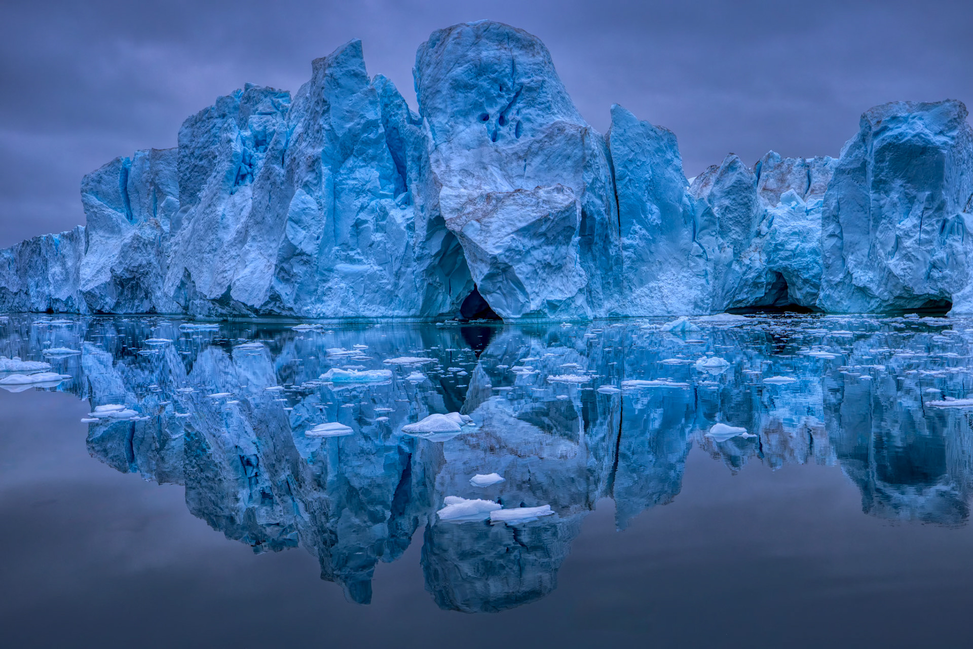 SCULPTED BY TIME  |  This monumental iceberg, photographed off the coast of Greenland, looks less like floating ice and more like an ancient fortress rising from the sea. Towering walls, deep crevasses, hidden caverns, and jagged spires reveal centuries of compression within Greenland's massive ice sheet before this giant finally broke free and began its journey through Arctic waters.The perfectly calm sea transforms the scene into a natural mirror, doubling the iceberg's scale and creating an almost surreal sense of symmetry. Small fragments of ice scattered across the foreground hint at the constant process of change taking place. Even a structure of this immense size is slowly being reshaped by wind, waves, and time.Photographed during the soft blue light of an Arctic evening, the image captures the raw grandeur of Greenland's frozen wilderness. The intricate textures, glowing blue tones, and cathedral-like formations evoke a sense of awe that is difficult to describe.