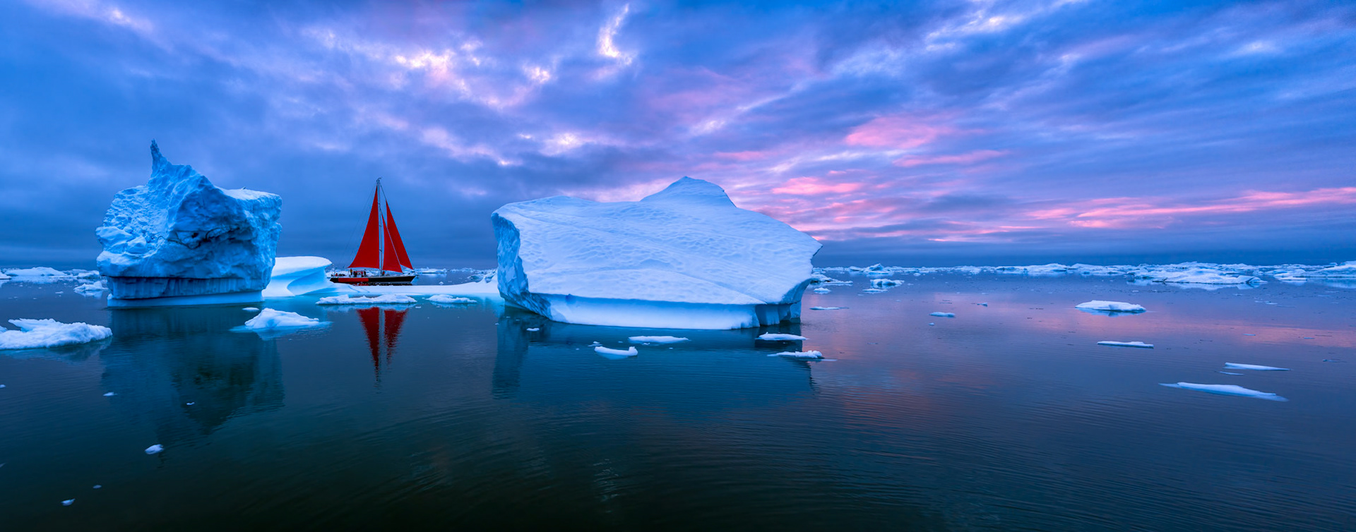 CHINESE DRAGON  |  A brilliant red sailboat threads its way through a frozen maze of Greenlandic icebergs, bringing a splash of color to an otherwise blue and white world. Surrounded by ancient ice and calm Arctic waters, the vessel appears almost suspended in time, dwarfed by the immense scale of the landscape around it.Photographed during the soft light of a Greenland summer evening, the scene captures a rare balance of adventure and tranquility. Massive icebergs, some centuries old, drift silently across the sea while streaks of pink and lavender spread across the sky. The vivid red sails create a striking focal point, guiding the eye through a wilderness where nature still reigns supreme. It is a reminder that even in one of the most remote places on Earth, beauty often reveals itself in the contrast between the immense power of nature and the small human stories unfolding within it.