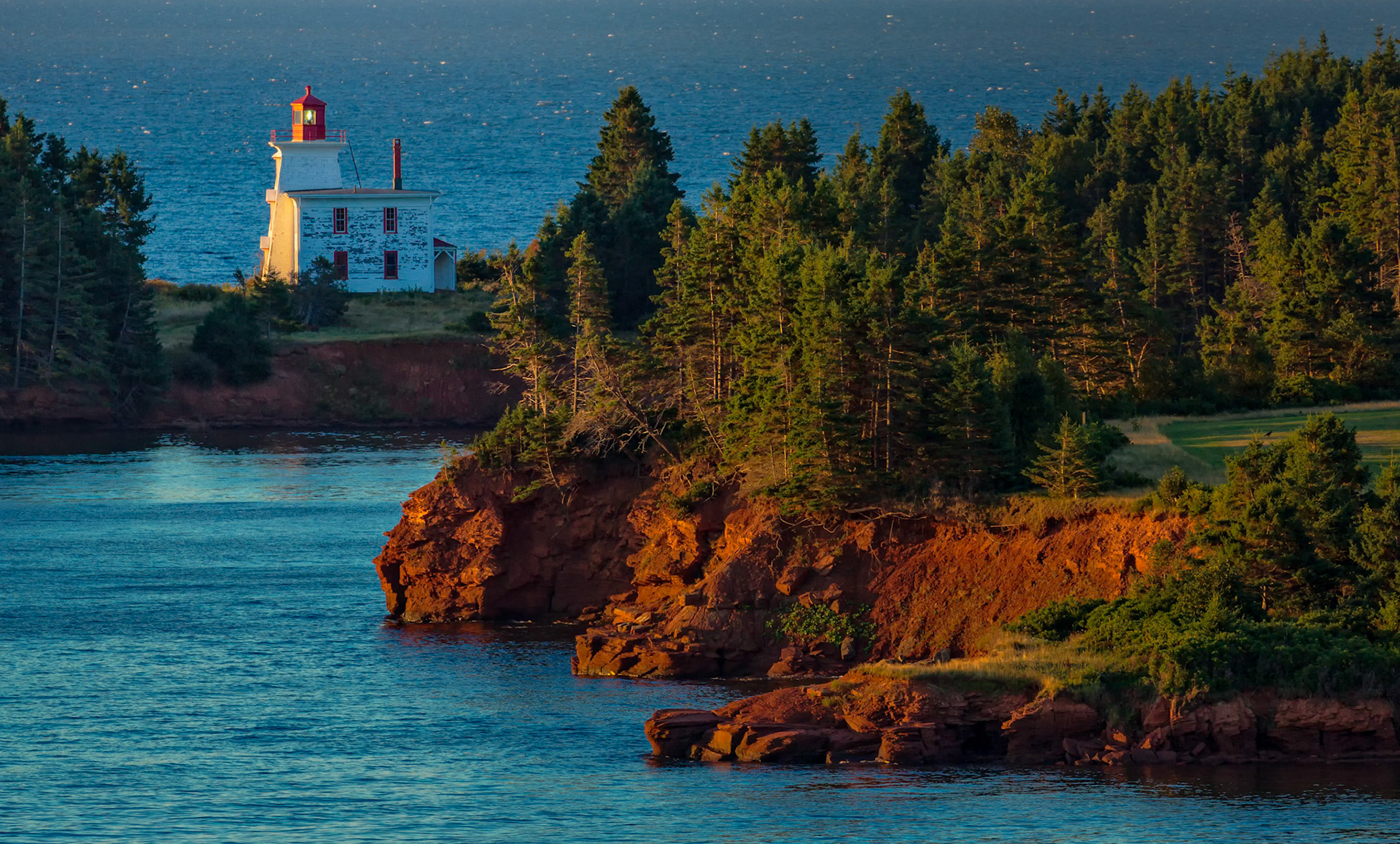 KEEPER OF THE RED CLIFFS  |  Perched above the rugged shoreline of Nova Scotia, the lighthouse stands watch over waters that have shaped generations of sailors, fishermen, and coastal communities. Bathed in the warm glow of autumn light, it serves as both landmark and guardian, a quiet symbol of resilience along Canada's eastern shore.The fiery red cliffs, sculpted by wind and sea, contrast beautifully with the deep blue water and evergreen forests that blanket the coastline. Together they create a landscape that feels both wild and welcoming, where the rhythm of the ocean has long defined life on the land.Autumn brings a special magic to this corner of Nova Scotia. The lower angle of the sun enriches every color, illuminating the shoreline with a warmth that hints at the coming winter. In this peaceful moment, the lighthouse becomes more than a structure. It becomes a beacon of permanence, standing steadfast between forest, sea, and sky.