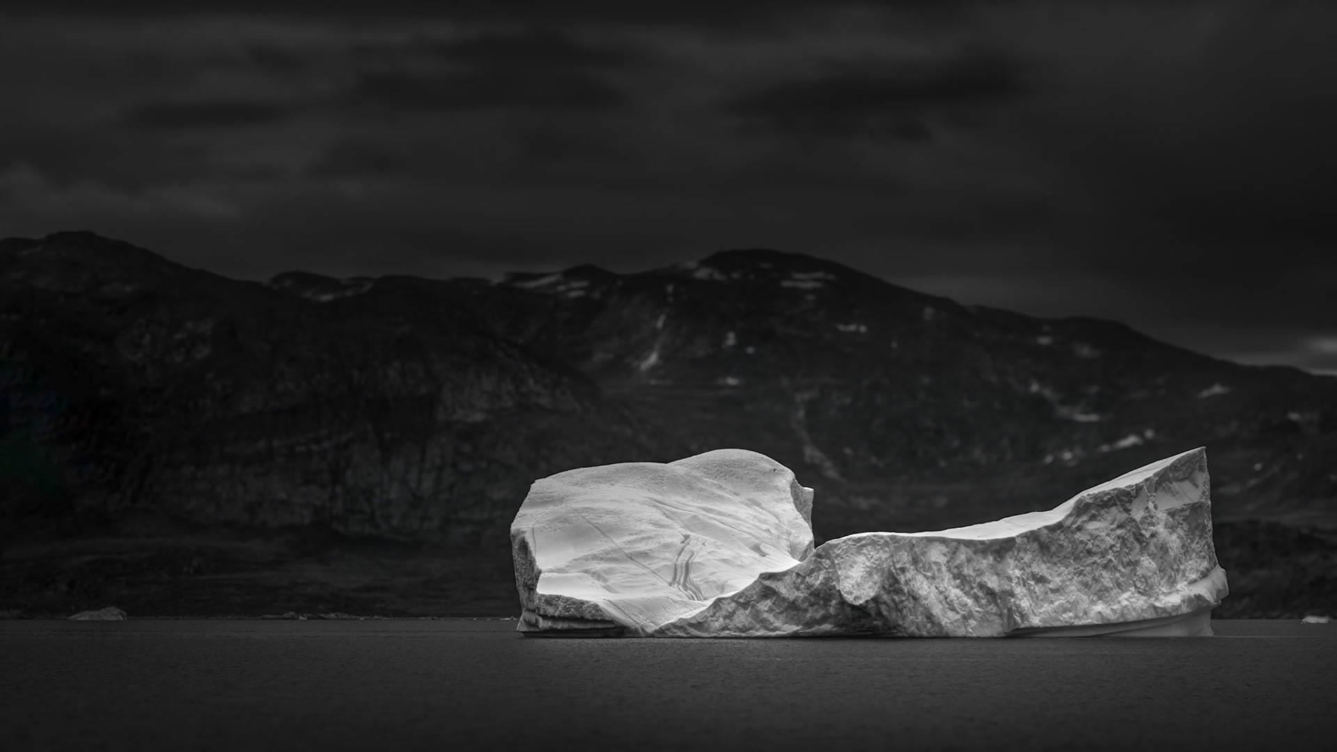 THE SHAPE OF SILENCE  |  Adrift beneath Greenland's towering coastal mountains, this massive iceberg seems to glow against the dark landscape behind it. Its sculpted surfaces reveal centuries of compressed snow and ice, transformed by time, pressure, and the slow forces of nature into a floating work of art.What captivated me was the contrast. The iceberg appears almost luminous against the shadowed cliffs, drawing the eye to its intricate textures, subtle curves, and delicate bands etched into the ice. Despite its immense size, there is a quiet elegance to its form.Moments like this inspire both awe and humility. These frozen giants may drift silently through Arctic waters for years, yet their existence is fleeting. This image captures a brief encounter with one of nature's most extraordinary creations, a reminder of both the beauty and impermanence of the polar world.