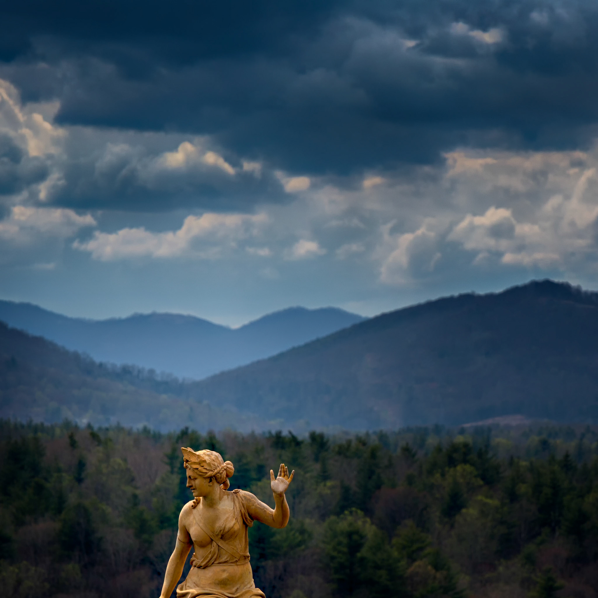 SENTINEL OF THE BLUE RIDGE  |  A garden statue stands against the backdrop of the Blue Ridge Mountains as storm clouds gather overhead. Layers of ridges recede into the distance while shafts of light break through the clouds, adding depth and atmosphere to the scene.The image is less about the statue itself and more about its relationship to the landscape. Positioned against the vast mountain view and dramatic sky, the figure provides a human scale that emphasizes the size and power of the approaching weather.