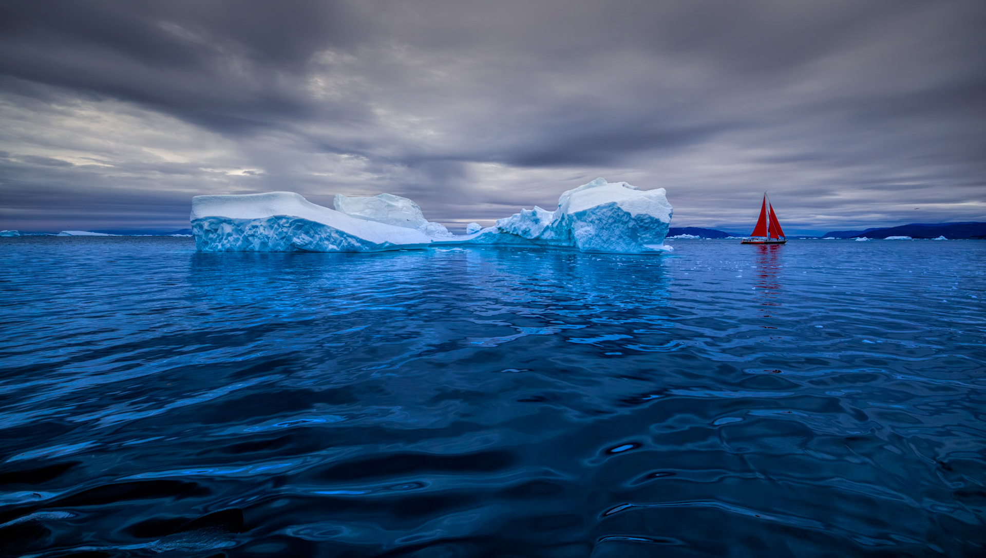 EDGE OF THE QUIET EARTH  |  The Arctic has a way of challenging every assumption. Standing on the deck among drifting icebergs and water so impossibly blue it seemed illuminated from within, I found myself staring into the sea and thinking the unthinkable: I wanted to jump in. The water was only a few degrees above freezing, yet its clarity and color were irresistible, as if the ocean itself was inviting exploration.Photographed along Greenland's rugged coast, the scene captures the contrast that defines the Arctic. Ancient icebergs, born from glaciers centuries ago, drift silently beneath a brooding sky while a lone sailboat with vivid red sails brings a touch of warmth and humanity to the vast wilderness. The dark, rolling water fills the foreground, drawing the viewer into a landscape that feels untouched by time. It is a place where beauty is so overwhelming that, for a moment, even the most inhospitable waters on Earth seem inviting.