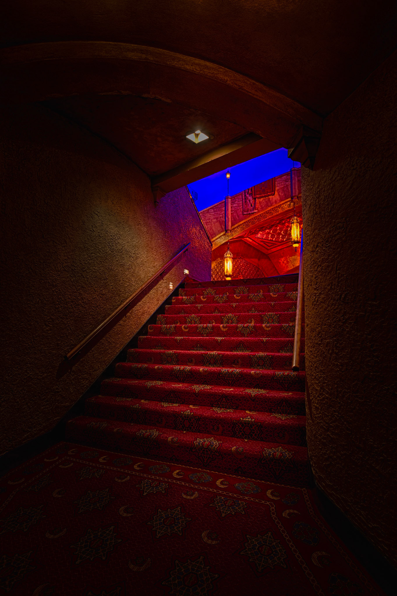 INTO THE HOUSE  |  A narrow staircase leads from the shadows toward the warm glow of the Fox Theatre beyond. Richly patterned carpet, hanging lanterns, and hints of ornate architecture create a sense of anticipation, as though each step brings visitors closer to another era.The composition uses light and color to guide the eye upward. Dark walls frame the scene, while the vivid blue ceiling and warm amber lighting reveal just enough of the theater's Moorish inspired design to spark curiosity about what lies ahead.For generations of theatergoers, moments like this have been part of the Fox experience. Before the performance begins, before the curtain rises, there is the journey through the building itself, where every hallway, stairway, and archway contributes to the sense of occasion.