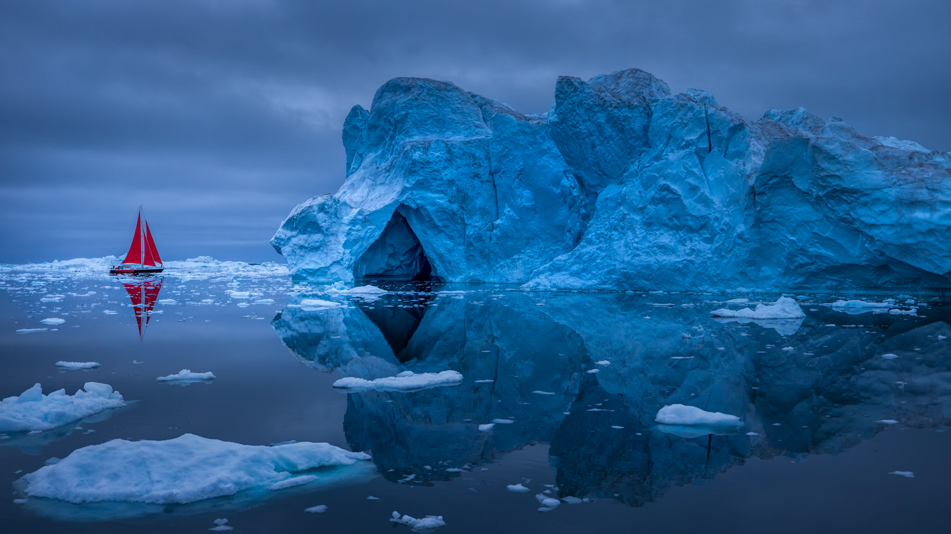 CRIMSON PASSAGE  |  A lone sailboat with brilliant red sails drifts through the icy waters of Greenland, providing a striking contrast to the towering blue iceberg that dominates the scene. The immense wall of ice rises from the sea like a frozen cathedral, its sculpted surfaces and cavernous opening revealing centuries of compression, fracturing, and transformation within Greenland's vast ice sheet.The perfectly calm water reflects both ice and sky, creating a sense of stillness rarely experienced in such a wild environment. The small vessel offers a powerful sense of scale, reminding viewers that what appears impressive in a photograph is, in reality, enormous beyond imagination. Photographed during the soft light of an Arctic summer evening, this image captures the wonder of exploring one of the most remote regions on Earth, where ancient ice, endless horizons, and moments of profound solitude combine to create an unforgettable experience.