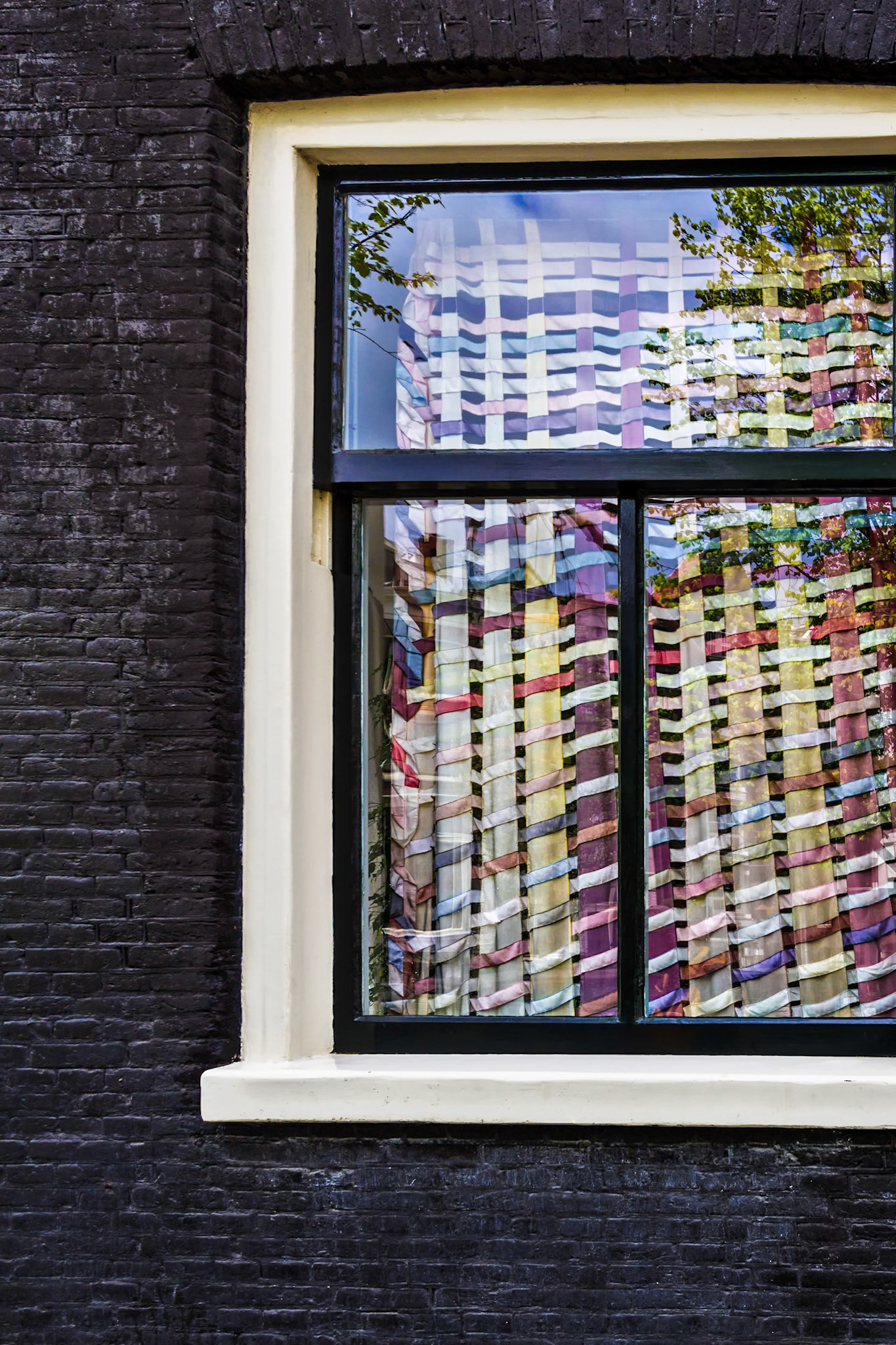 BEHIND THE WINDOW  |  I discovered this scene while wandering through Amsterdam, where even an ordinary window can become a canvas. The colorful woven fabric hanging inside caught my attention, but what made the photograph for me was the reflection of the trees and sky layered across it. The result felt less like looking through a window and more like looking into two worlds at once.One of the things I enjoy most about street photography is finding these unexpected visual collisions. Nothing here was staged. The fabric, the reflection, and the old brick façade simply came together for a brief moment to create something far more interesting than any one element on its own.Amsterdam is known for its canals and architecture, but sometimes the city's character reveals itself in the smallest details hiding between the landmarks.