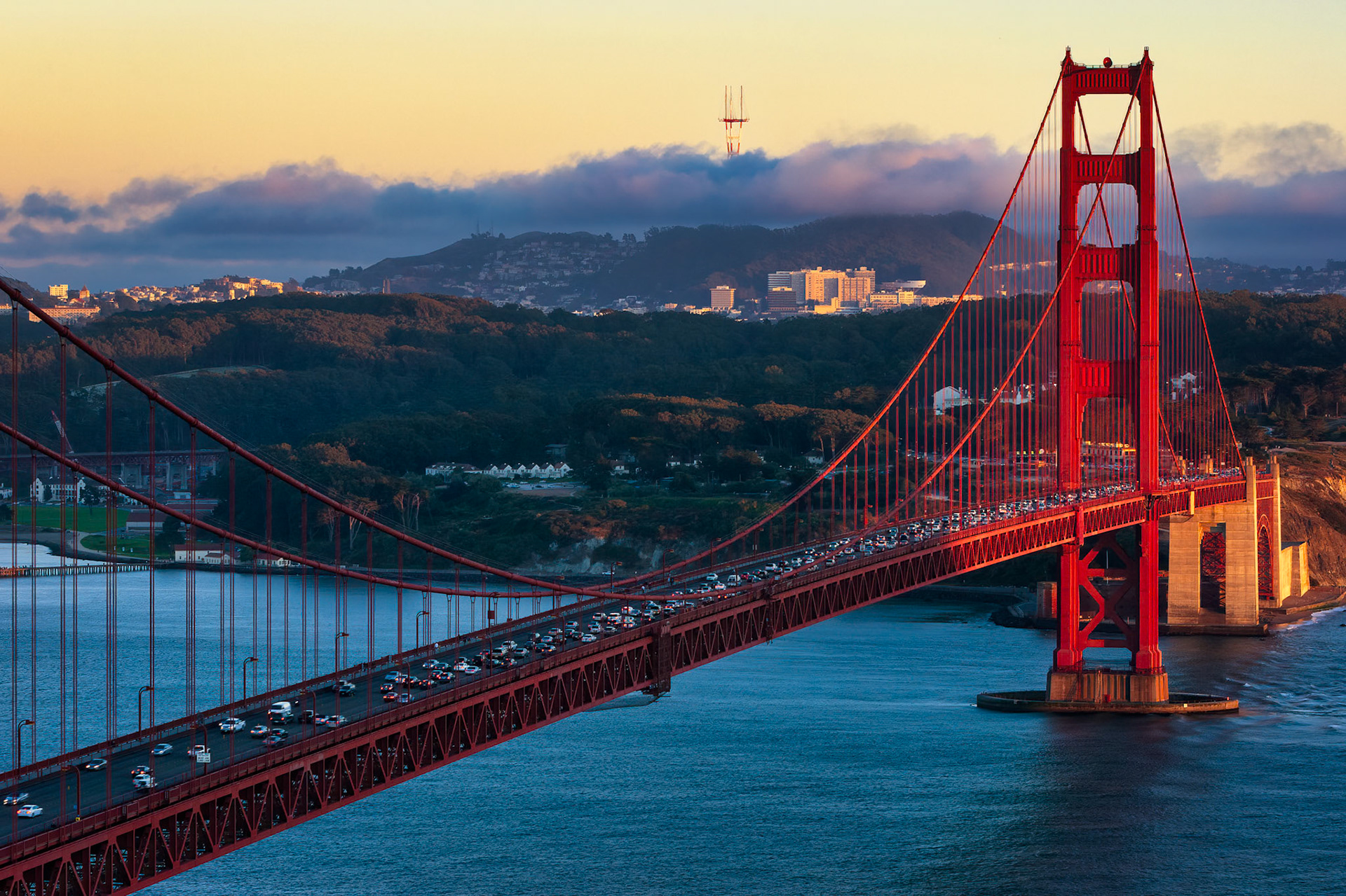 GOLDEN GATE BRIDGE FROM MARIN HEADLANDS | SAN FRANCISCO  |  Early morning light illuminates the Golden Gate Bridge as it spans the entrance to San Francisco Bay. The bridge's iconic International Orange towers stand in striking contrast to the cool blue water and the rolling hills beyond.Low clouds drift across the ridgeline behind the bridge, while the first sunlight of the day highlights portions of the surrounding landscape. The steady stream of vehicles crossing the span serves as a reminder that, beyond its status as a global landmark, the bridge remains an essential transportation link for the region.The elevated perspective showcases both the engineering elegance of the suspension bridge and its dramatic setting between the city and the Pacific Ocean, capturing a familiar landmark in the quiet transition from night to day.