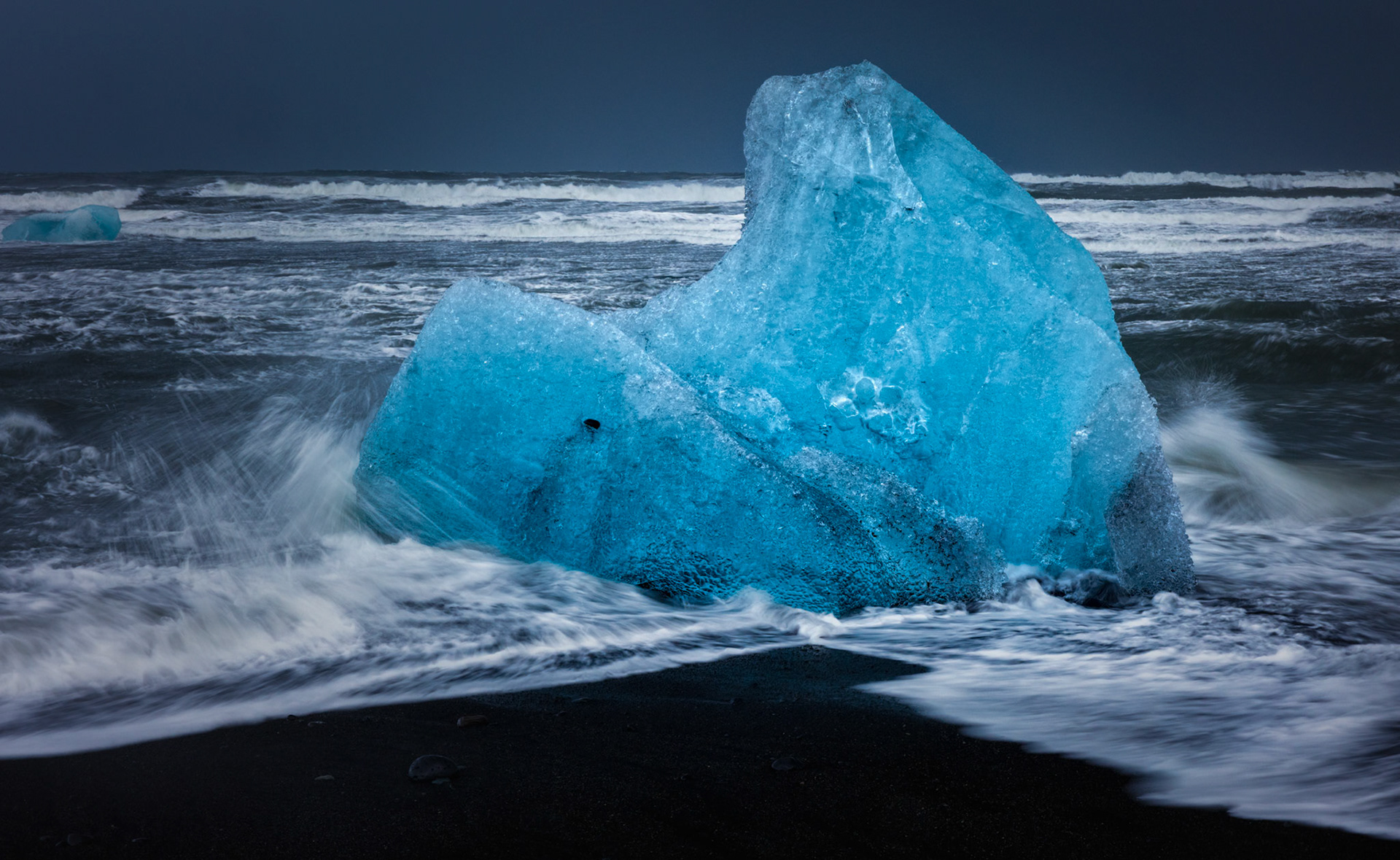 MIDNIGHT SAPPHIRE  |  Along Iceland's Diamond Beach, glacier ice begins its final journey. After breaking free from a glacier and drifting through the lagoon, these ancient fragments are carried into the North Atlantic, where powerful waves relentlessly reshape them before they ultimately disappear into the sea.What drew me to this piece was its resilience. The ice stood firm against the crashing surf, glowing with an almost electric blue intensity against the black volcanic sand. Every wave seemed determined to claim it, yet for this brief moment it remained defiant, sculpted into a form that looked both fragile and enduring.This image captures the meeting of opposites: ice and fire, permanence and impermanence, calm beauty and raw power. The glacier ice may have taken centuries to form, but its final days are measured in hours and tides, making every encounter along Diamond Beach a fleeting gift.
