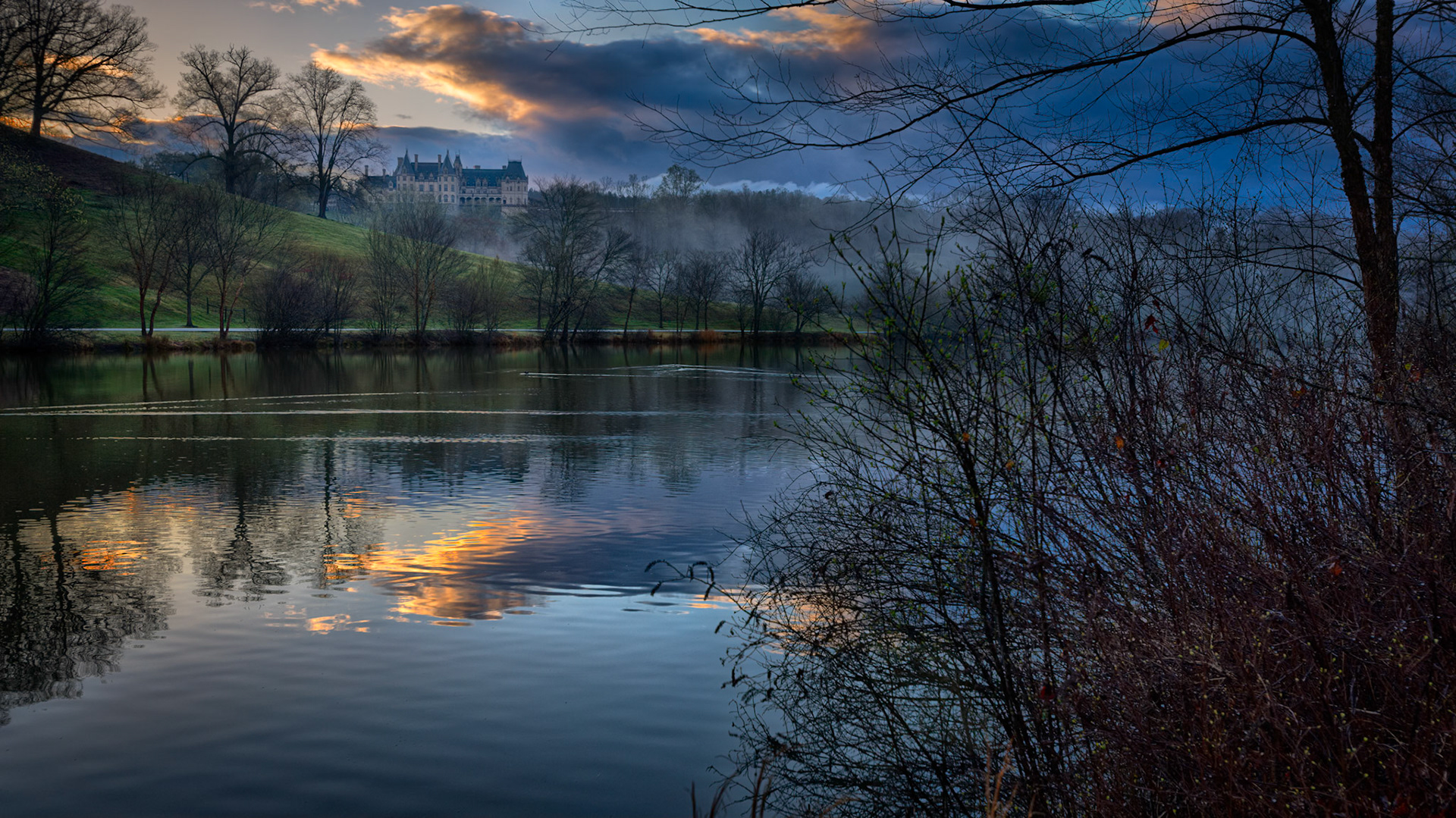 MORNING AT THE ESTATE  |  The Biltmore Estate sits quietly on a distant hillside as evening light breaks through the clouds and reflects across the lake. Mist lingers among the trees, softening the landscape and adding depth to the scene.Rather than dominating the image, the estate becomes part of the larger landscape. The water, reflections, and fading light work together to create a peaceful moment at the close of day.