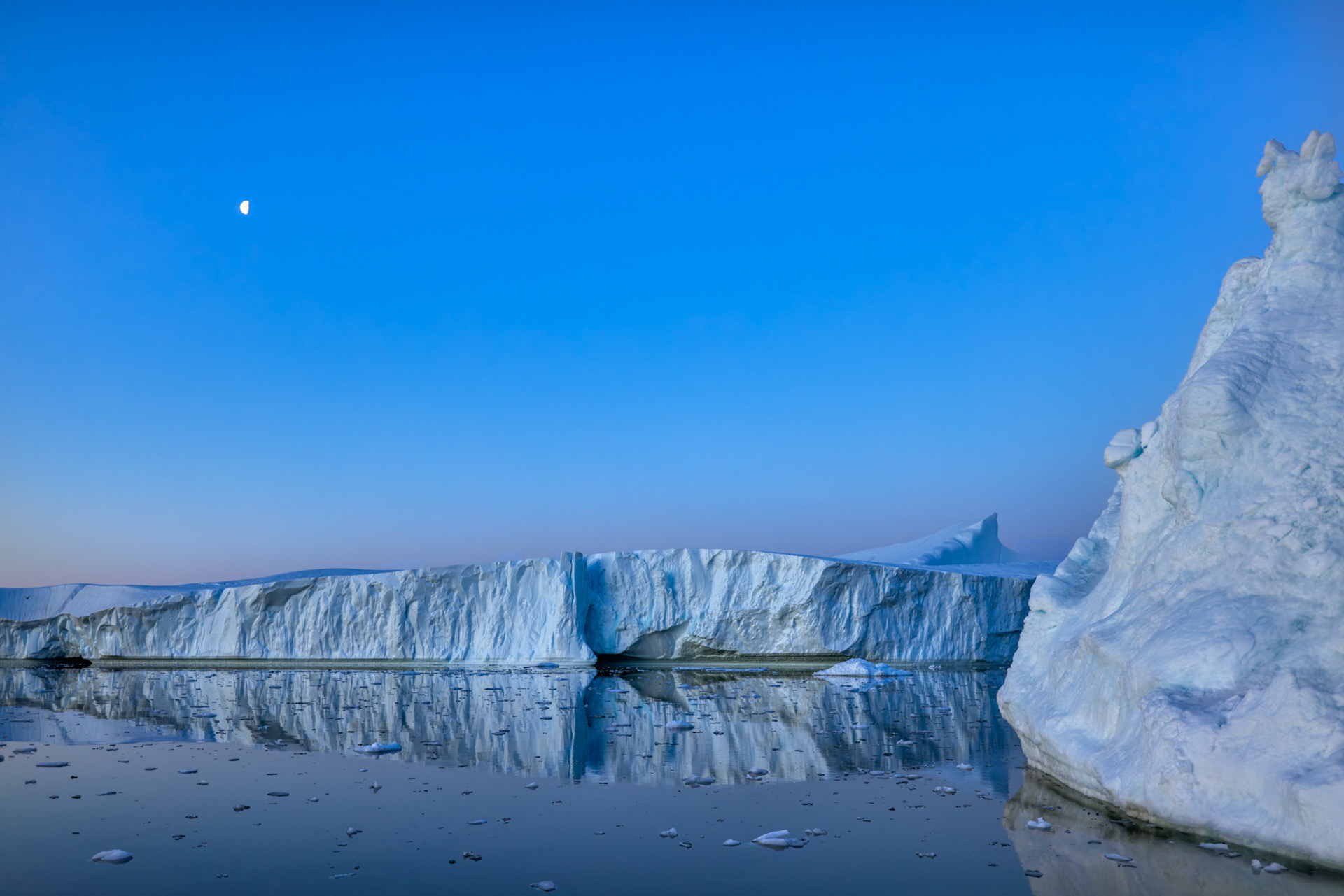 WHERE THE MOON RESTS  |  In the endless twilight of a Greenland summer, a lone moon hangs above a frozen landscape where time seems to stand still. Massive icebergs drift silently across glassy water, their reflections merging sky and sea into a world of soft blues and quiet serenity.What drew me to this scene was its simplicity. There is no dramatic sunrise or towering mountain, only the subtle beauty of the Arctic at rest. The moon, the ice, and the calm water create a sense of peace that is difficult to describe and impossible to forget.Moments like this remind me why I return to the polar regions. In a world filled with noise and motion, the Arctic offers something increasingly rare: silence, space, and the opportunity to simply be present.