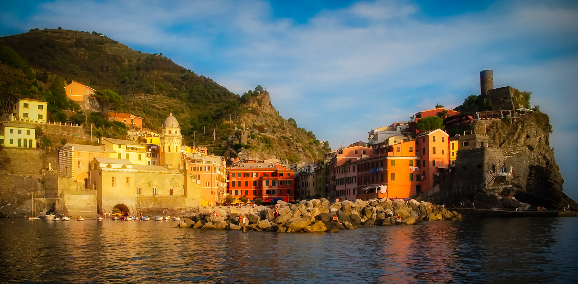 COLOR AGAINST THE SEA  |  As the last light of day sweeps across the Ligurian coast, Vernazza glows as though illuminated from within. Warm sunlight paints the pastel facades in shades of gold, amber, and coral, transforming this centuries old fishing village into a living watercolor.Approaching by sea offers a perspective few travelers experience. The village slowly reveals itself from behind the rocky headlands, its harbor sheltered between steep cliffs that have protected generations of fishermen, sailors, and dreamers. The ancient watchtower stands guard above the sea, a reminder of Vernazza's enduring relationship with the Mediterranean.In that fleeting moment before sunset fades, the village seems suspended between history and beauty. The gentle movement of the water, the warmth of the evening light, and the timeless architecture create a scene that feels less like a destination and more like a memory waiting to be revisited.