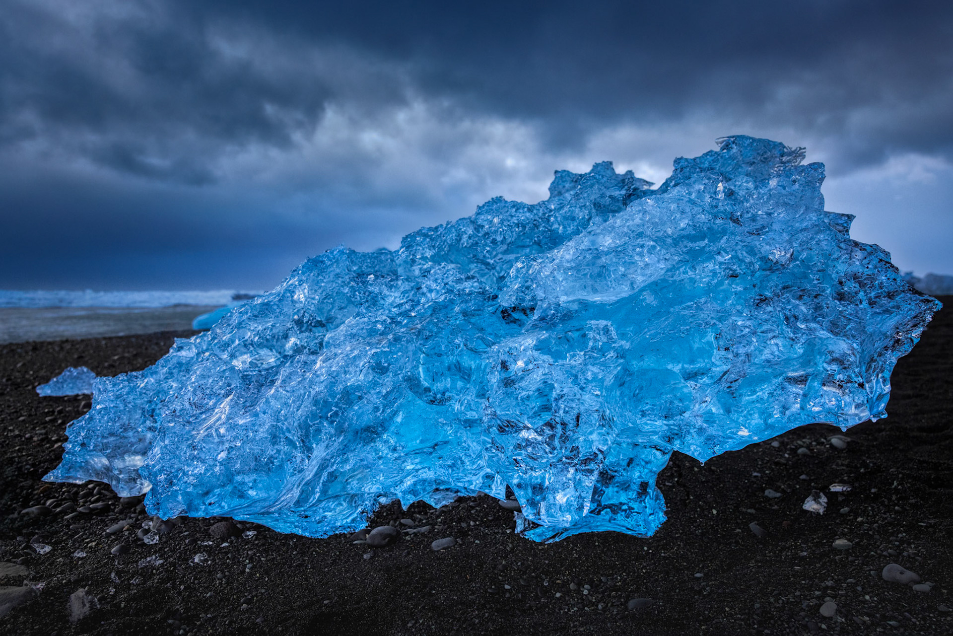 GLACIAL JEWEL  |  Along Iceland's famous Diamond Beach, fragments of ancient glacier ice are carried from the lagoon to the North Atlantic, where they wash ashore like precious gemstones scattered across black volcanic sand. Each piece is unique, shaped by years of compression within the glacier and sculpted further by wind, waves, and time.What fascinated me about this ice formation was its extraordinary clarity. Unlike ordinary ice, glacier ice has been compressed over centuries, forcing out air bubbles and allowing light to penetrate deep within it. The result is a brilliant blue glow that seems to originate from within the ice itself.Standing on the beach that day, the contrast felt almost surreal. Crystal blue ice resting on jet black sand beneath a brooding Icelandic sky created a scene that seemed more imagined than real. It was a reminder that some of nature's most beautiful creations are also among its most temporary, destined to melt back into the sea from which they came.