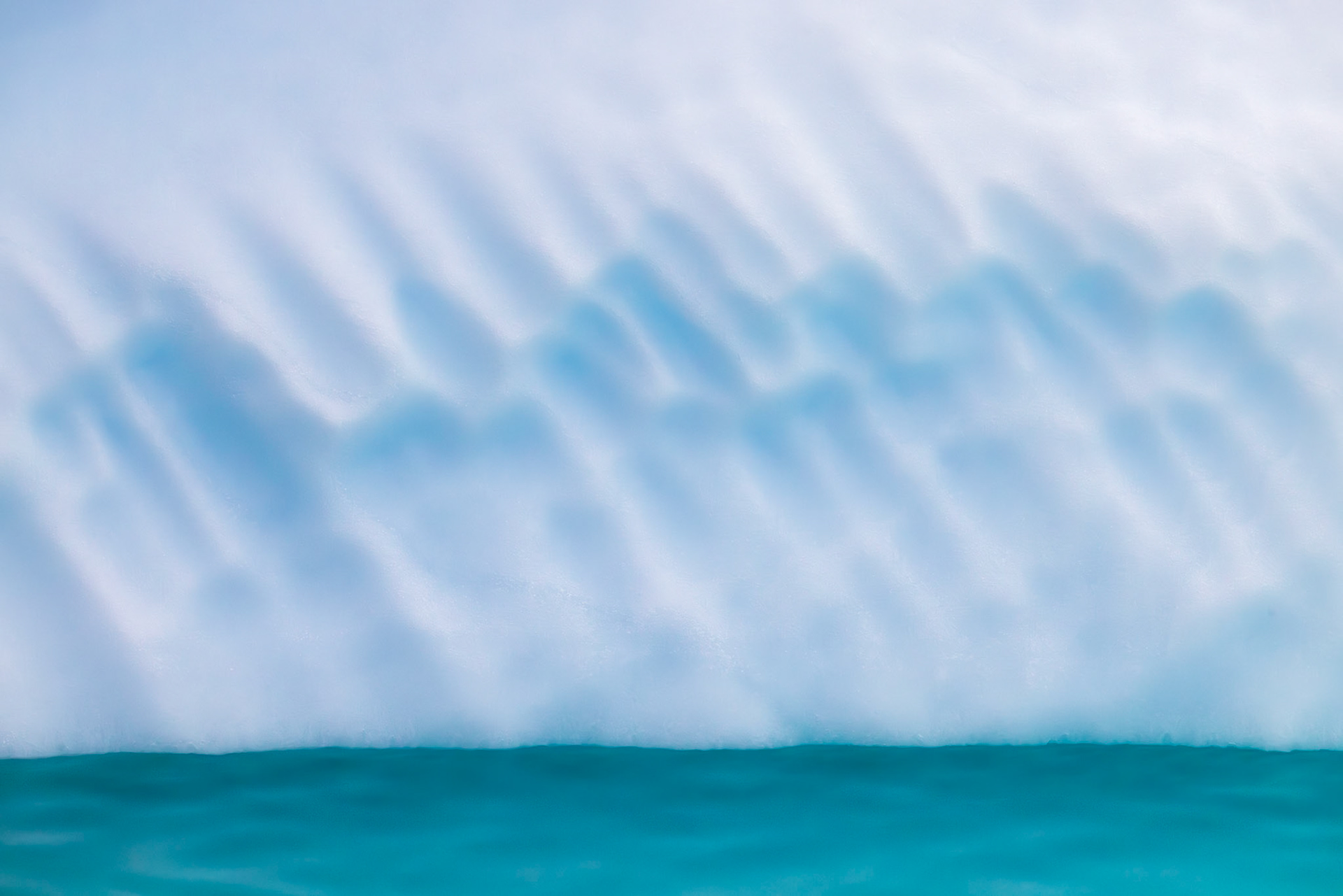 BETWEEN ICE AND SEA  |  At first glance, this image feels almost minimalist, yet it captures one of the most powerful forces in nature. The crisp white face of an iceberg rises from the turquoise waters of the North Atlantic, its surface sculpted by wind, waves, and time. Long blue shadows stretch across the ice, revealing subtle ridges and contours that might otherwise go unnoticed.Photographed in Greenland, this scene marks the meeting point between ancient glacial ice and the ever changing ocean. The iceberg may have spent centuries compressed within a glacier before finally breaking free and beginning its slow journey south. The simplicity of the composition draws attention to the remarkable contrast between the frozen stillness of the ice and the restless energy of the sea, creating an image that feels both tranquil and timeless.