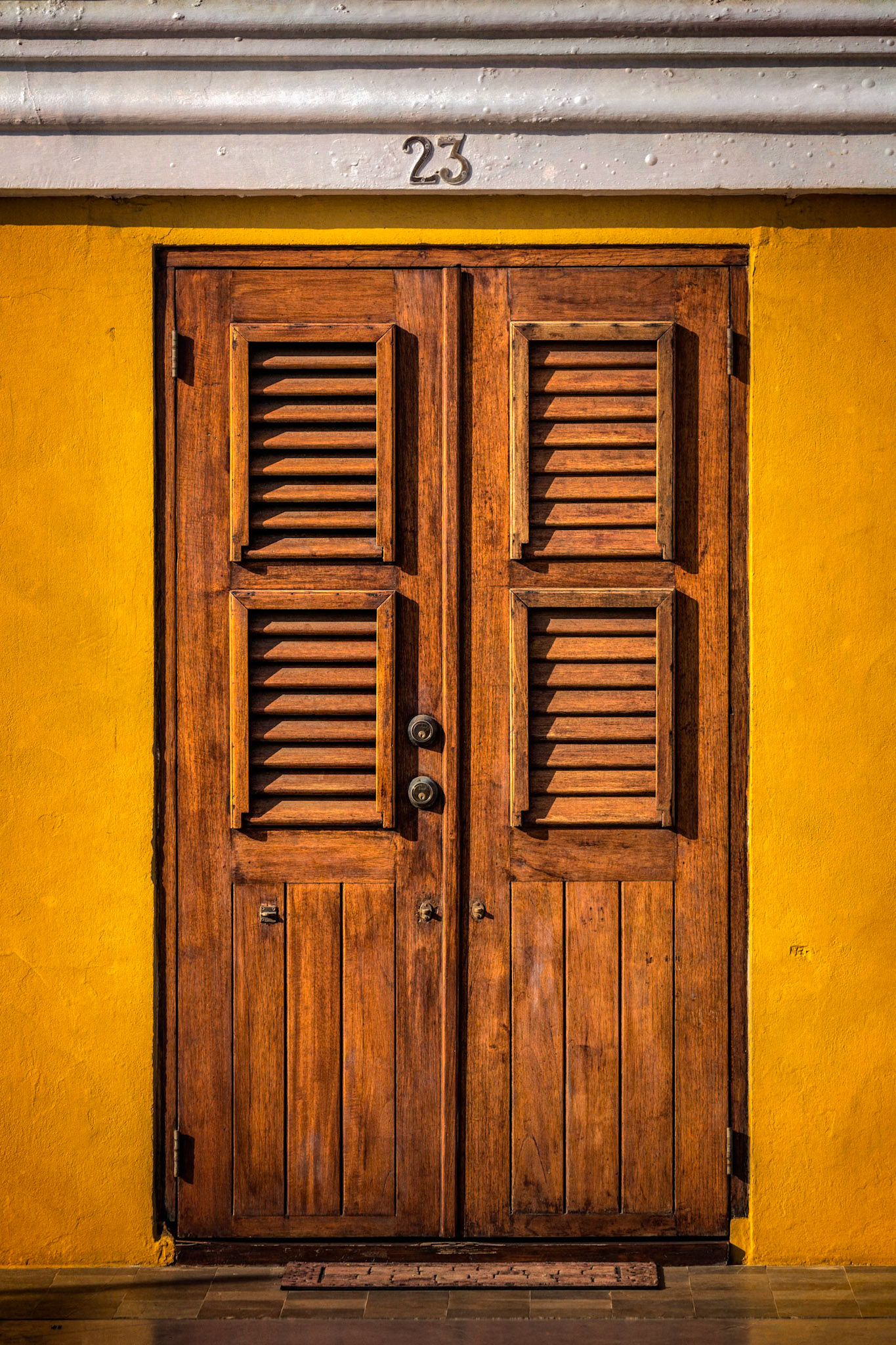 DOOR NUMBER 23  |  Travel often teaches us that beauty does not have to be grand to be memorable.Walking through Aruba, I found myself drawn not to a famous landmark or sweeping vista, but to a simple doorway. The warm yellow stucco, weathered wood, and soft Caribbean light combined to create a scene that felt timeless. There was nothing extraordinary about it, yet it stopped me in my tracks.What caught my eye was the harmony of the elements. The rich grain of the wooden shutters contrasted beautifully against the vibrant wall, while the symmetry of the door created a sense of balance and calm. Above it all, the number 23 quietly marked an address that could belong to anyone and everyone at the same time.Photographs like this remind me that every door carries a story. Behind it may be a family gathering for dinner, a couple beginning their day, or generations of memories woven into the walls. We rarely know the stories hidden beyond a closed door, and perhaps that mystery is part of their appeal.In a world often focused on dramatic destinations, I am continually reminded that some of the most rewarding photographs come from slowing down and noticing the details. Beauty is not always found in the extraordinary. Sometimes it is found in a doorway glowing in the afternoon sun.