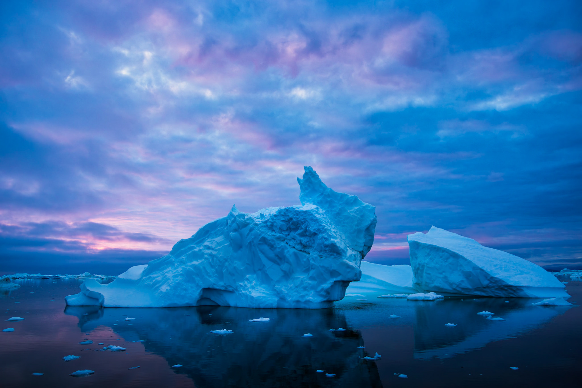 CATHEDRAL OF BLUE ICE  |  Bathed in the soft glow of an Arctic twilight, this iceberg drifts silently through the waters of Greenland as shades of blue, lavender, and pink blend across the sky. Sculpted by centuries of compression within a glacier, its jagged peaks and weathered surfaces tell a story that began long before modern civilization.The stillness of the water creates a near perfect reflection, blurring the boundary between ice and sky. In this remote corner of the Arctic, moments like these are fleeting. The light changes by the minute, the ice is constantly moving, and every iceberg exists only temporarily before wind, waves, and warming seas reshape it once again. The scene is both peaceful and humbling, a reminder of nature's immense beauty and the fragile permanence of even the largest creations on Earth.