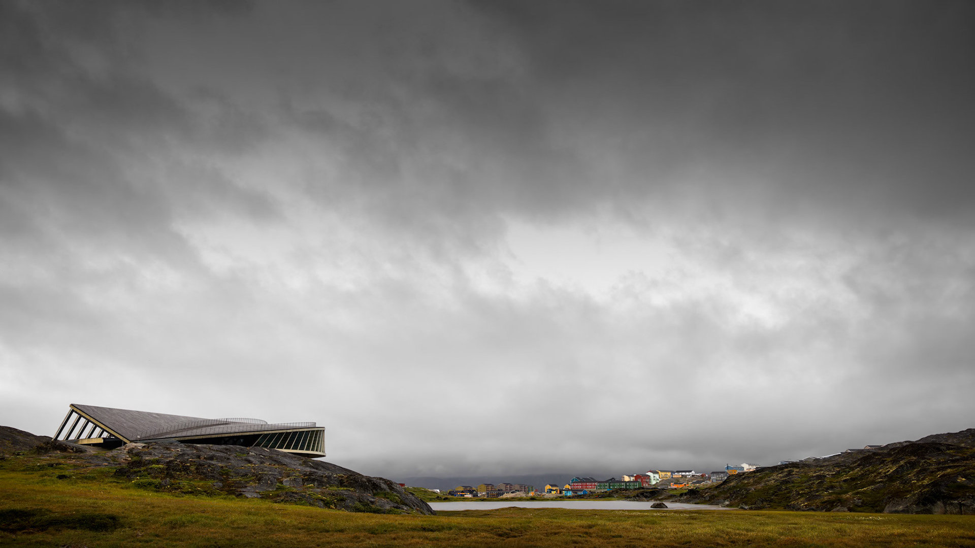 EDGE OF SETTLEMENT  |  Perched quietly on the rocky edge of Ilulissat, this striking modern structure shares the landscape with one of the most extraordinary natural wonders on Earth. Beyond it lies the Ilulissat Icefjord, a UNESCO World Heritage Site where ancient glaciers continue their slow journey toward the sea.What captivated me was the contrast. The sleek geometry of the building feels undeniably contemporary, yet it sits in complete harmony with a landscape shaped by ice, stone, and time. The colorful homes of Ilulissat appear in the distance, a reminder of the resilient community that calls this remote corner of Greenland home.The vast sky dominates the frame, creating a sense of scale that is difficult to comprehend until experienced firsthand. Beneath those heavy Arctic clouds, the land feels both intimate and immense. The building appears almost secondary, humbled by the environment surrounding it.This image speaks to humanity's desire to understand and connect with the natural world. Here, architecture does not compete with nature. It quietly observes it.