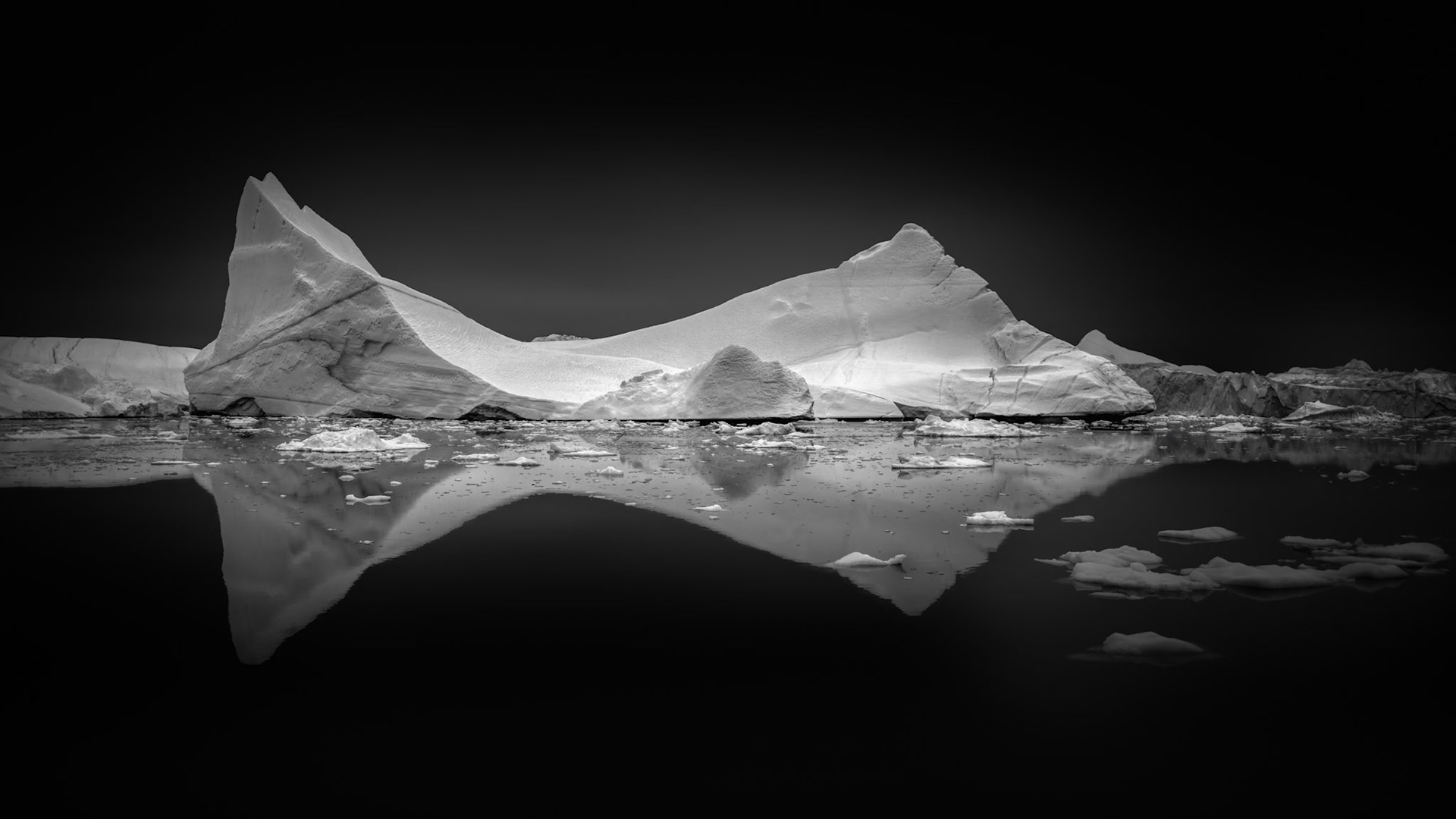 HELD AGAINST THE COLD  |  This iceberg, photographed along the coast of Greenland, takes on an almost sculptural quality in black and white. Stripped of color, the image draws attention to the elegant lines, textures, and immense scale of the ice itself. Its sweeping form and perfect reflection create a striking sense of balance, transforming a fleeting moment in nature into something timeless.  The iceberg may have spent centuries locked within Greenland's vast ice sheet before finally breaking free and beginning its slow journey toward the North Atlantic. In the still water, its reflection doubles the composition, creating the illusion of a frozen mountain suspended between two worlds. The monochromatic treatment emphasizes the quiet solitude of the Arctic, where silence, light, and ice combine to create scenes of extraordinary simplicity and beauty. Far from civilization, this ancient traveler drifts through one of Earth's last great wildernesses.