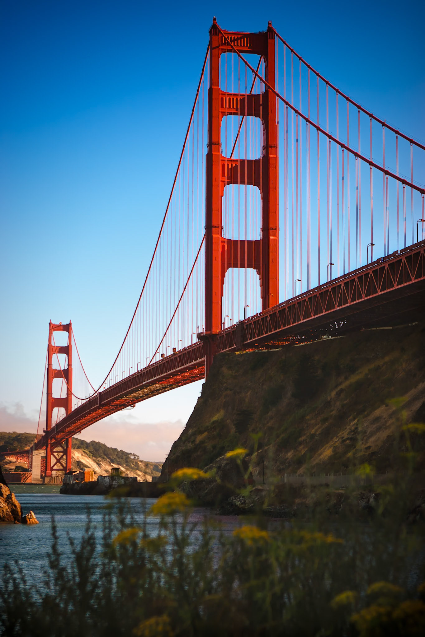 GOLDEN GATE BRIDGE FROM SAUSALITO | SAN FRANCISCO  |  The Golden Gate Bridge spans the entrance to San Francisco Bay, connecting the city to Marin County across the waters of the Golden Gate Strait. Photographed from below the northern approach, the composition emphasizes the bridge's soaring towers, sweeping suspension cables, and distinctive International Orange color.The low vantage point accentuates the scale of the structure while the surrounding cliffs and shoreline provide context for its dramatic setting. Soft foreground vegetation adds depth and a natural frame without competing for attention.Completed in 1937, the Golden Gate Bridge remains one of the most celebrated engineering achievements in the world and an enduring symbol of San Francisco. This perspective highlights both its monumental presence and the elegance of its design.
