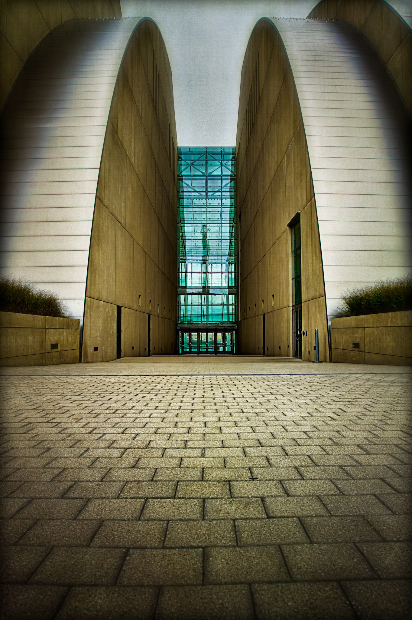 A PATH TO PERFORMANCE  |  The sweeping arches of the Kauffman Center frame a narrow corridor that draws the eye directly to the glass entrance beyond. Standing low to the ground emphasized the repeating pattern of the pavers and strengthened the symmetry of the scene.What caught my attention was the contrast between the massive curved walls and the delicate grid of glass at the center. The architecture feels both monumental and inviting, guiding visitors toward the heart of the building.