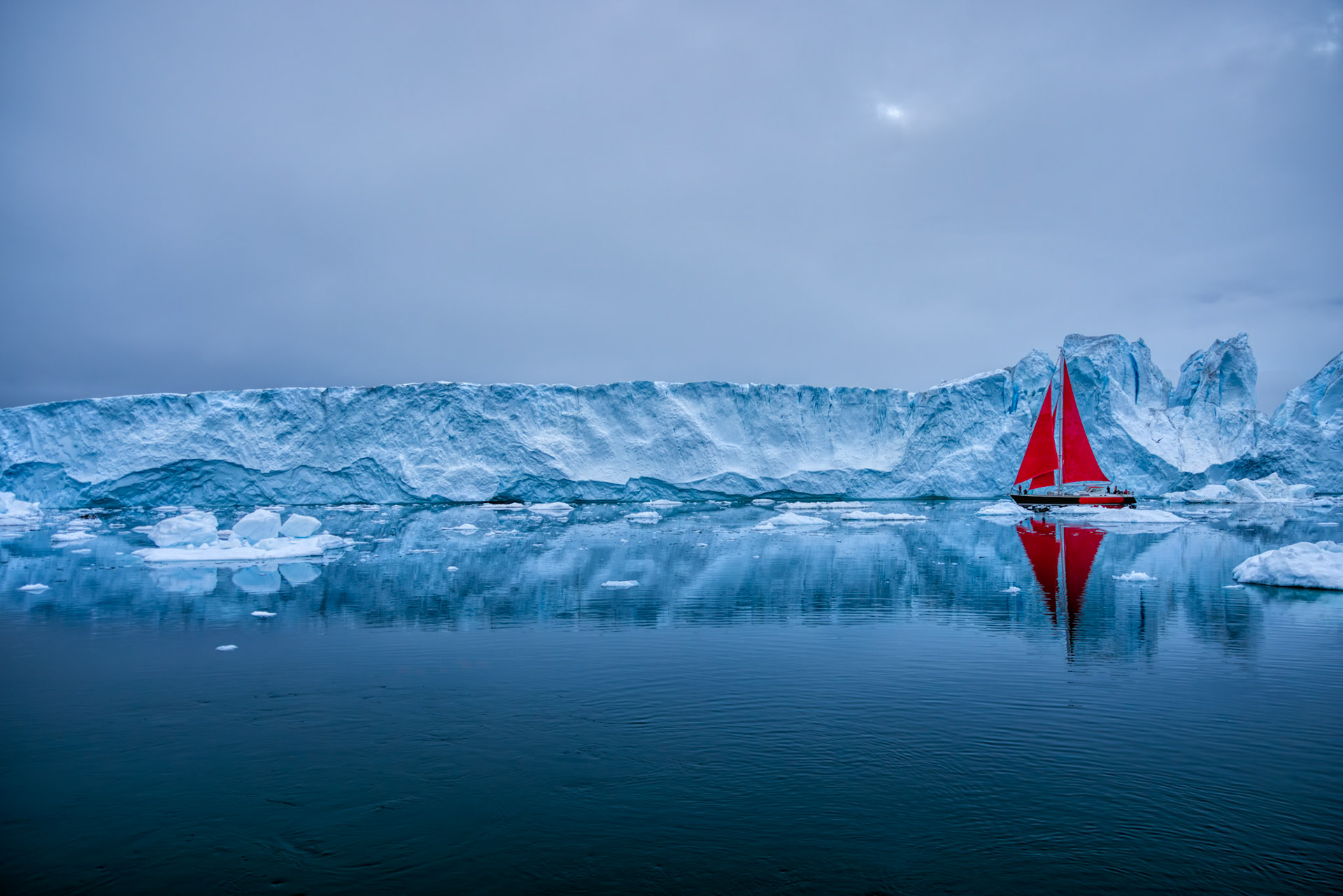 EDGE OF STILLNESS  |  A vivid red sailboat drifts across the still waters of Greenland, dwarfed by a towering wall of ancient ice stretching across the horizon. The contrast is striking. The brilliant sails provide a sense of scale, revealing just how immense these icebergs truly are. What appears from a distance to be a frozen cliff is actually a fragment of a glacier that may have spent hundreds, even thousands, of years compressed within Greenland's vast ice sheet before breaking free into the sea.  The calm water mirrors both ice and sky, creating a scene of remarkable serenity despite the immense forces at work. Icebergs of this size are constantly changing, slowly melting, rolling, and fracturing as they continue their journey through Arctic waters. Moments like this are rare. The combination of perfect reflections, soft polar light, and a lone vessel navigating among these frozen giants transforms the landscape into something that feels almost otherworldly, a reminder of the scale and beauty that still