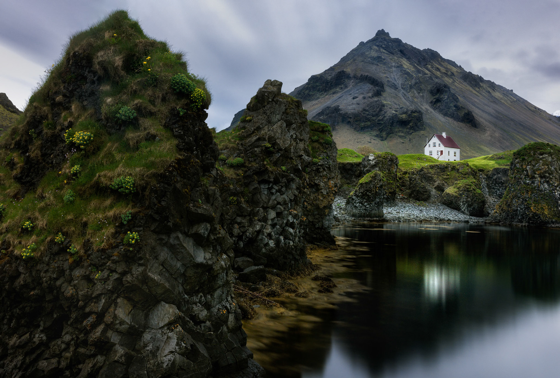 THE HOUSE BENEATH THE MOUNTAIN  |  Nestled beneath the towering slopes of Iceland's Snæfellsnes Peninsula, a solitary farmhouse stands surrounded by volcanic cliffs, weathered rock formations, and the still waters of a hidden cove. The scene feels almost untouched by time, where nature remains the dominant presence and human habitation is little more than a quiet footnote.