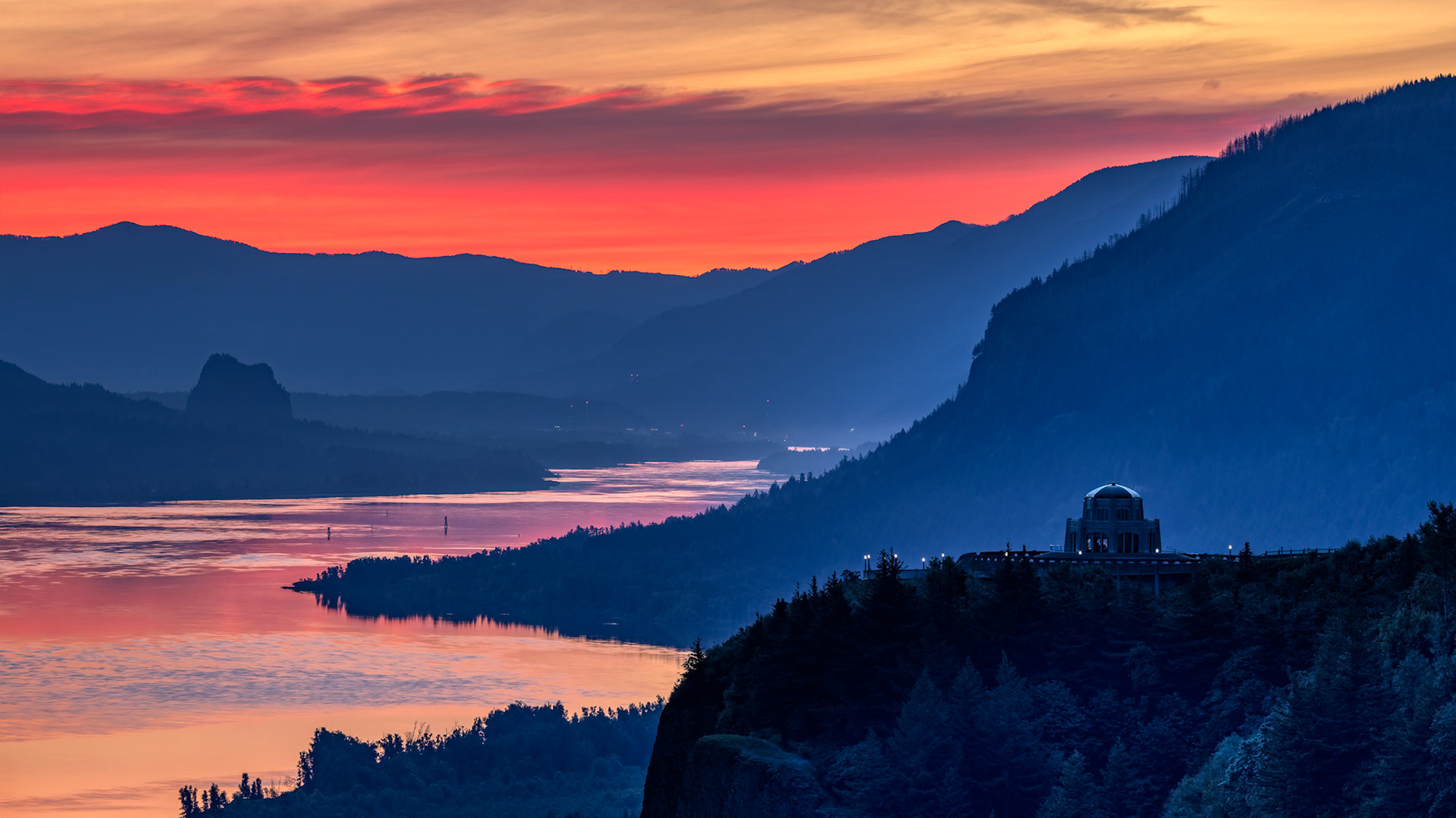 COLUMBIA AT DAWN  |  Perched high above the Columbia River Gorge, the historic vista house overlooks one of the Pacific Northwest's most spectacular landscapes. As the last light of day ignites the horizon, the river winds through layers of blue mountains, reflecting the colors of a fading sunset.The scene balances grandeur and serenity. The sweeping gorge conveys a sense of vastness, while the illuminated landmark anchors the landscape with a touch of human history. Together they create a view that has inspired travelers for generations.This image captures that fleeting moment between day and night when the sky glows with color, the river becomes a ribbon of light, and the Columbia Gorge reveals its timeless beauty.