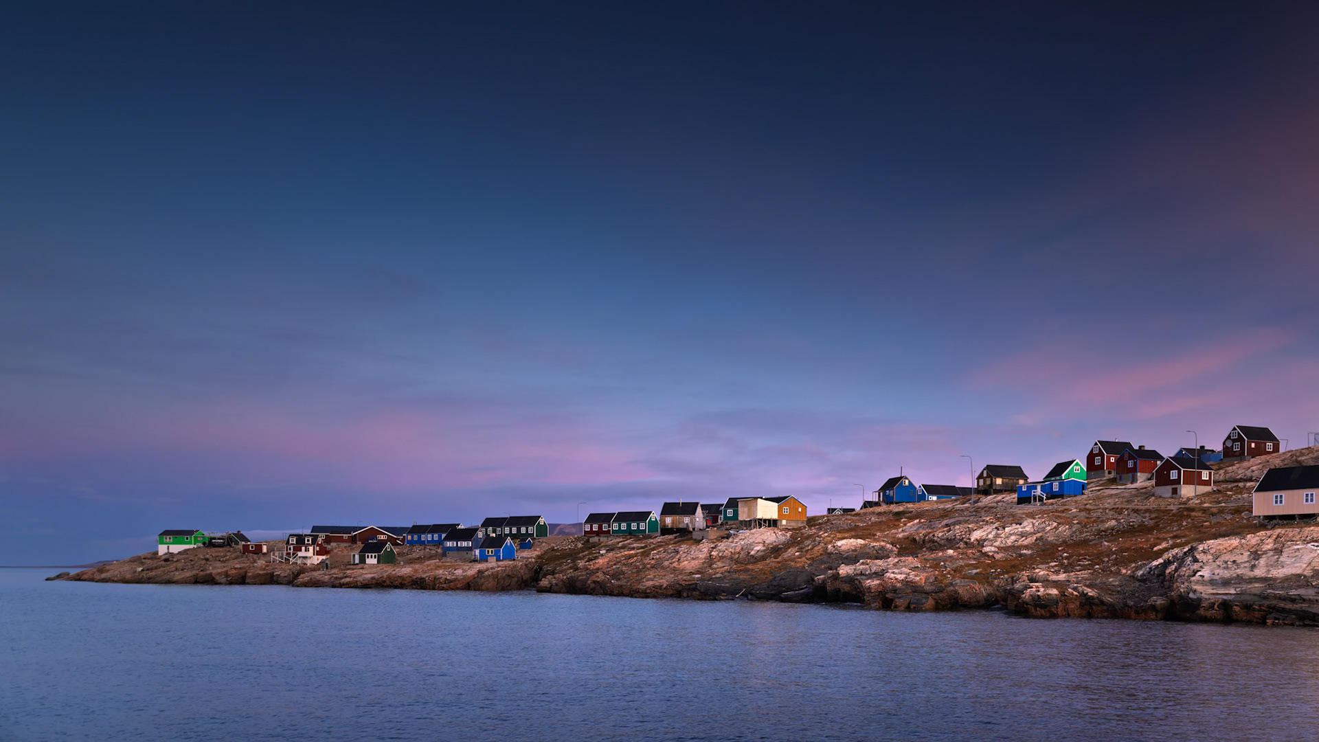 TWILIGHT IN ITTOQQORTOORMIIT  |  Soft Arctic twilight settles over the colorful homes of Ittoqqortoormiit, Greenland, as the village stretches along the rocky shoreline beneath a violet sky. The quiet water and fading light give the scene a sense of isolation, warmth, and calm at the edge of Scoresby Sound.