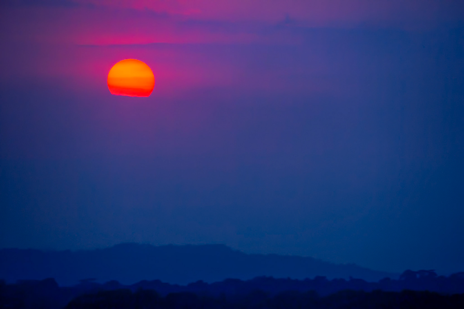 THE NIGHT LETS GO  |  As dawn breaks over the Panama Canal, the rising sun emerges through layers of tropical haze, transforming the sky into a delicate blend of violet, indigo, and crimson. The landscape below remains in shadow, waiting patiently as the first light of a new day spreads across the horizon.The simplicity of the scene is its strength. A solitary sun suspended above distant hills becomes the entire story, drawing attention to the quiet beauty that exists in the moments between darkness and daylight. The soft atmospheric layers create depth and mystery, blurring the boundary between earth and sky.There is a sense of anticipation in every sunrise, but especially here. For centuries, travelers have crossed this narrow passage between oceans, each journey beginning with a horizon much like this one. In the stillness of dawn, before the movement of ships and the activity of the day, the canal reveals a rare moment of calm, where light, color, and silence come together in perfect balance.