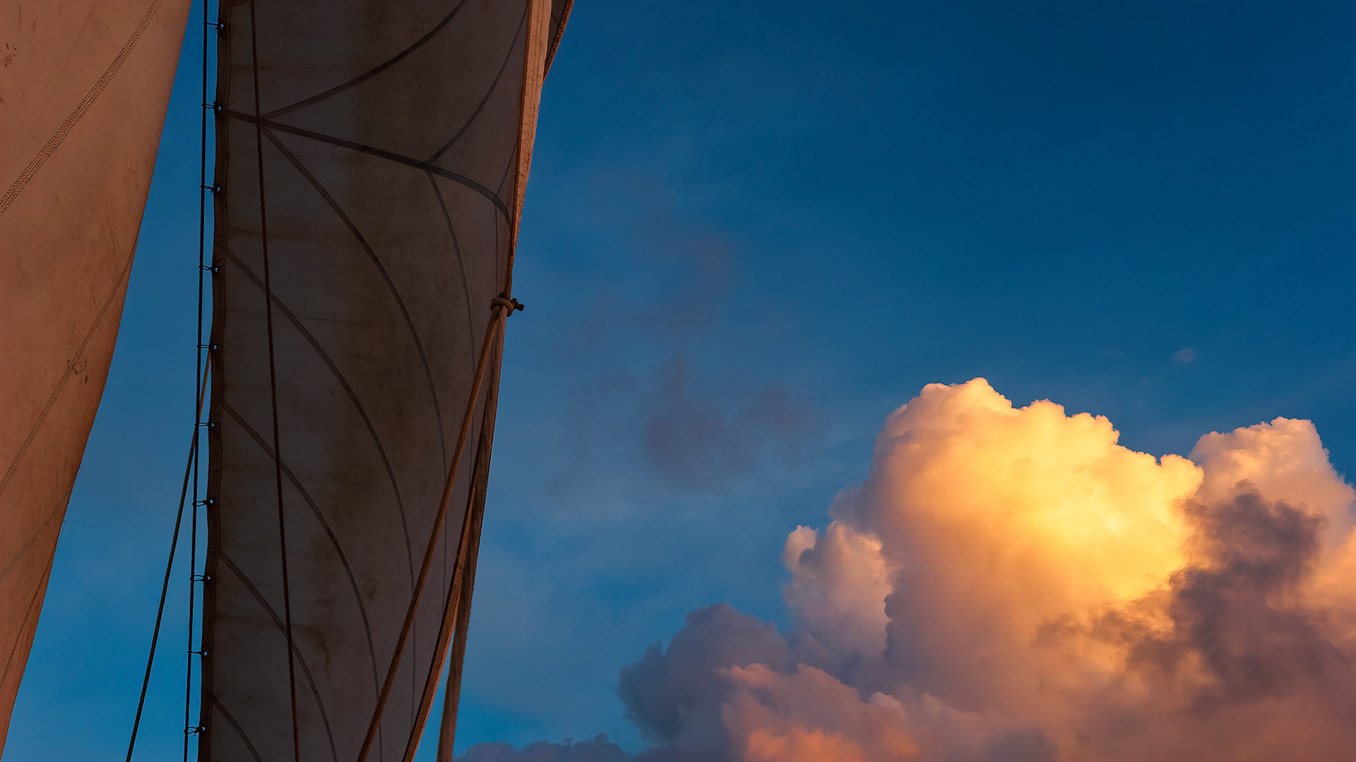 SAILS AND SUMMER SKY  |  As evening settles over the Caribbean, a towering cloud catches the last light of the sun, glowing like a lantern against the deepening blue sky. Beside it, the sail rises in quiet partnership with the wind, carrying the vessel steadily onward across open water.There is something timeless about traveling under canvas. The absence of engines allows every detail to emerge. The whisper of wind through the rigging, the gentle pull of the sails, and the changing colors of the sky become part of the journey itself.For a brief moment, sea, sky, and sail exist in perfect harmony. The illuminated cloud becomes both destination and companion, reminding us that some of life's most memorable passages are not measured by miles traveled, but by the beauty encountered along the way.