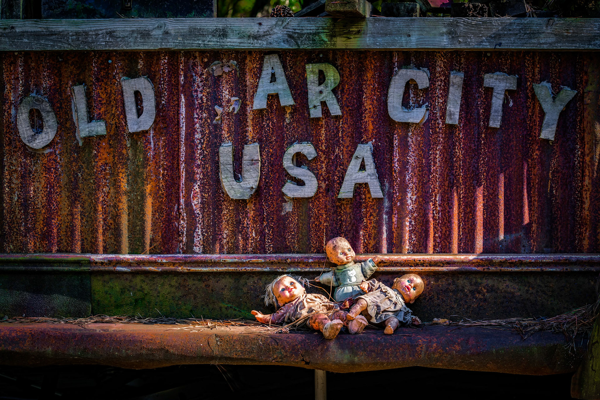 OLD CAR CITY, USA  |  This image captures the wonderfully strange spirit that makes Old Car City unlike any other automotive graveyard. A weathered corrugated metal sign boldly proclaims "OLD CAR CITY USA," its handmade letters standing against a backdrop of rust and age. Yet it is the unexpected detail below that transforms the scene from simple documentation into storytelling.Three abandoned dolls rest at the base of the sign, worn by years of exposure to the elements. Their faded faces and tattered clothing create a haunting juxtaposition against the rusted metal surrounding them. What might initially seem unsettling soon becomes oddly poignant. Like the forgotten automobiles scattered throughout the property, these dolls have become artifacts of another time, preserved through neglect rather than intention.