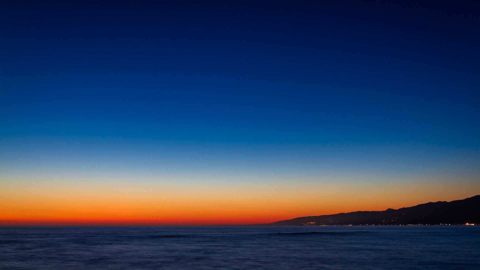 AFTER THE SUN  |  The day quietly surrenders to night along the California coast as a ribbon of amber light lingers above the horizon. Looking north from Santa Monica Beach, the ocean becomes a vast expanse of calm, reflecting the final colors of a sunset that seems reluctant to fade.The distant silhouette of the coastline stretches into the darkness, its scattered lights offering a gentle reminder of the millions of lives unfolding beyond the shoreline. Above, an immense gradient of color transitions from fiery orange to deep indigo, creating a sense of scale that makes both land and sea feel wonderfully small.There is no dramatic wave, no towering cliff, no obvious focal point. Instead, the beauty lies in simplicity. The meeting of water, sky, and fading light creates a moment of quiet reflection, where the world seems to slow down and the horizon becomes an invitation to simply pause and look.