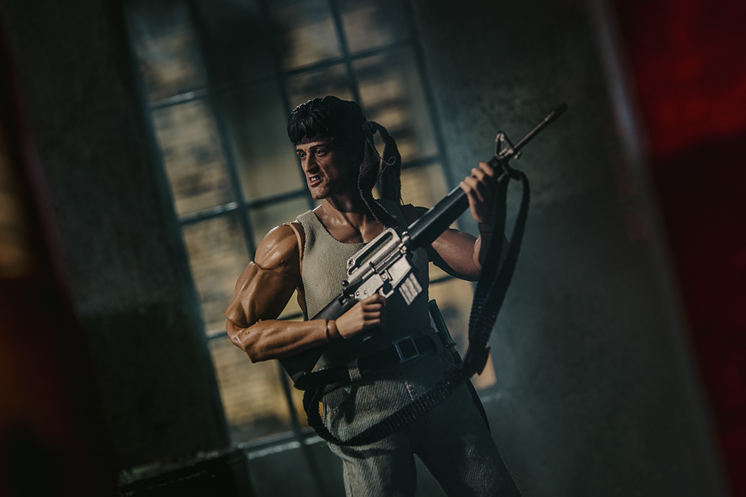 Na zdjęciu z filmu "Rambo: Pierwsza krew", widzimy Sylvestra Stallone w roli Johna Rambo, ikonicznej postaci w wojskowym stylu. Rambo ma długie, zmierzwione włosy, przepaskę na czole i ubrany jest w wojskową kurtkę lub kamizelkę. Jego twarz jest zabrudzona, z wyrazem skupienia i determinacji, co odzwierciedla jego gotowość do walki o przetrwanie. W jednej ręce trzyma duży, charakterystyczny nóż bojowy, a w tle możemy dostrzec gęsty las lub surowe górskie otoczenie, które tworzy dziką i nieprzyjazną atmosferę. Zdjęcie oddaje surowość i intensywność postaci, podkreślając jej samotność i siłę w obliczu trudnych wyzwań.