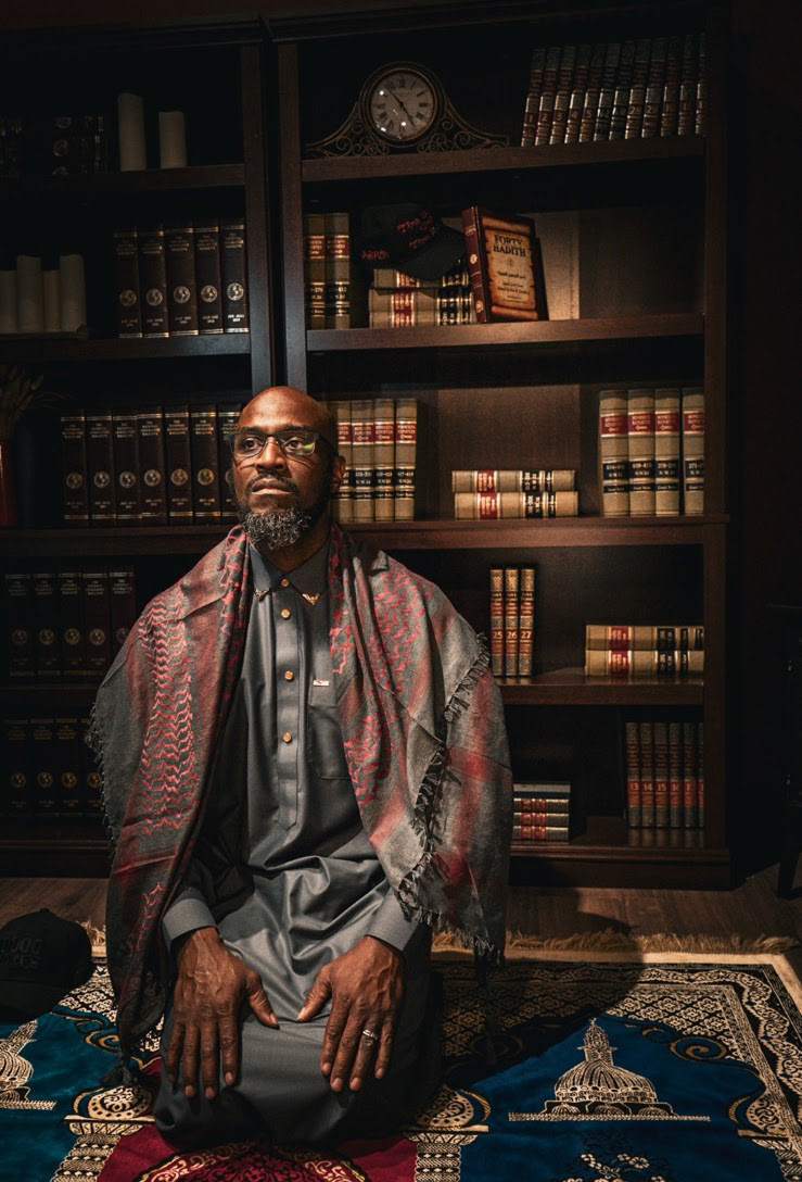 A man kneeling on a prayer rug in front of a book shelf featuring the book Forty Hadith