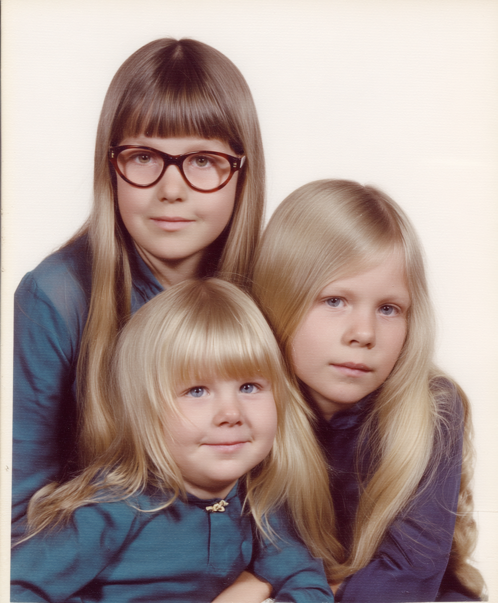 Eigene Darstellung (2025). Meine Mutter mit ihren Schwestern. prompt: family photo of three girls (4,6,8), one with horn-rimmed glasses, two with long blonde hair and round face, both resemble each other, two like twins, the oldest is dark-haired and has a narrow face, narrow nose and looks different, oldest looks different, she looks like a strange child, analog, 70s, west germany, dortmund, white background, only onw of the blonde girls is smiling,