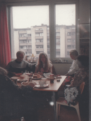 Eigene Darstellung (2025). Familienschnappschuss Wohnzimmer, schwangeres Mädchen mit Familie[KI-generiertes Bild]. Erstellt mit Midjourney. Prompt: “In a small living room of a 70s high-rise building in West Germany, 15 year old woman with long blond hair is heavily pregnant, she is not smiling, several old Polish men and women are sitting at the living room table Snapshot, coffe and lots of cake, flash, slightly blurred”