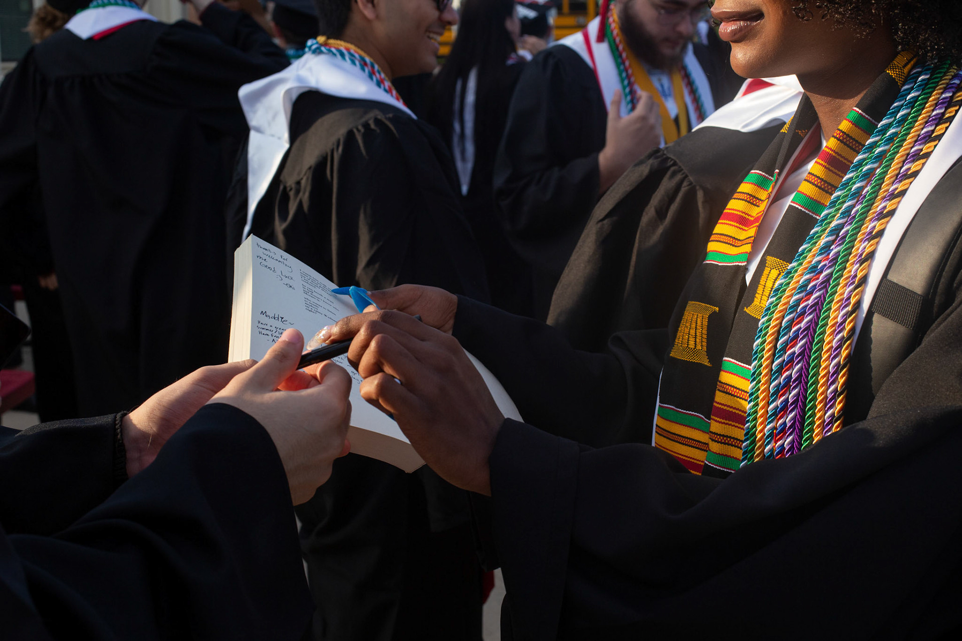 Topher Malone hands her classmates a copy of “Love is a Dog from Hell” to sign as a yearbook as they wait to load buses to graduation on May 26, 2023, on the Austin Community College Round Rock campus.
