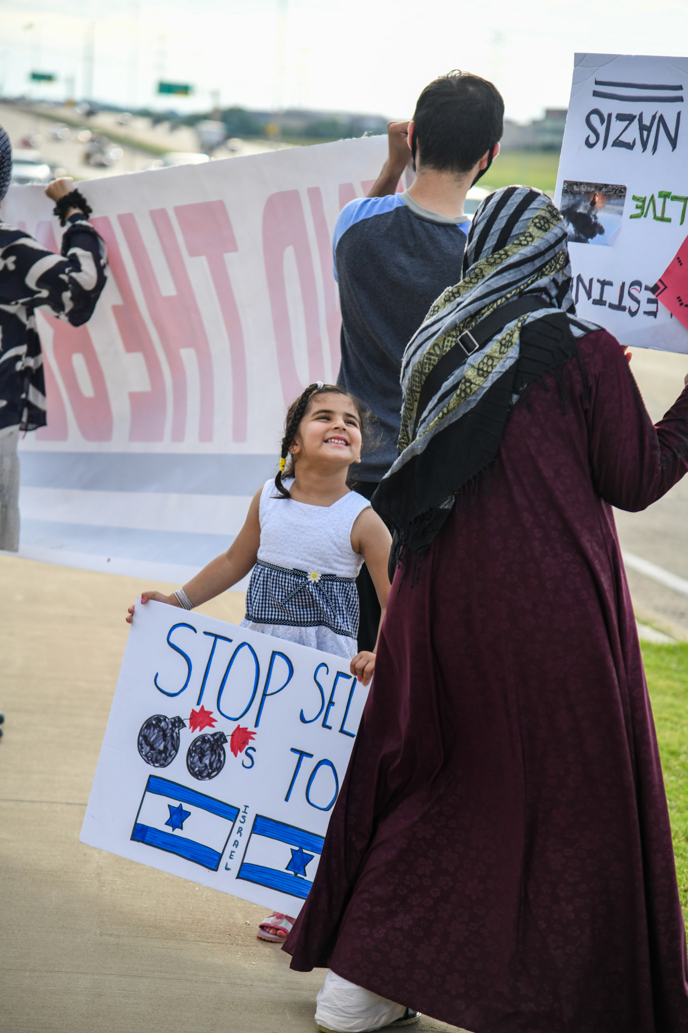 A young girl smiles while holding a sign reading "Stop selling [bombs] to [israel]" outside of Raytheon on June 4, 2021. Shot for The Dallas Morning News.
