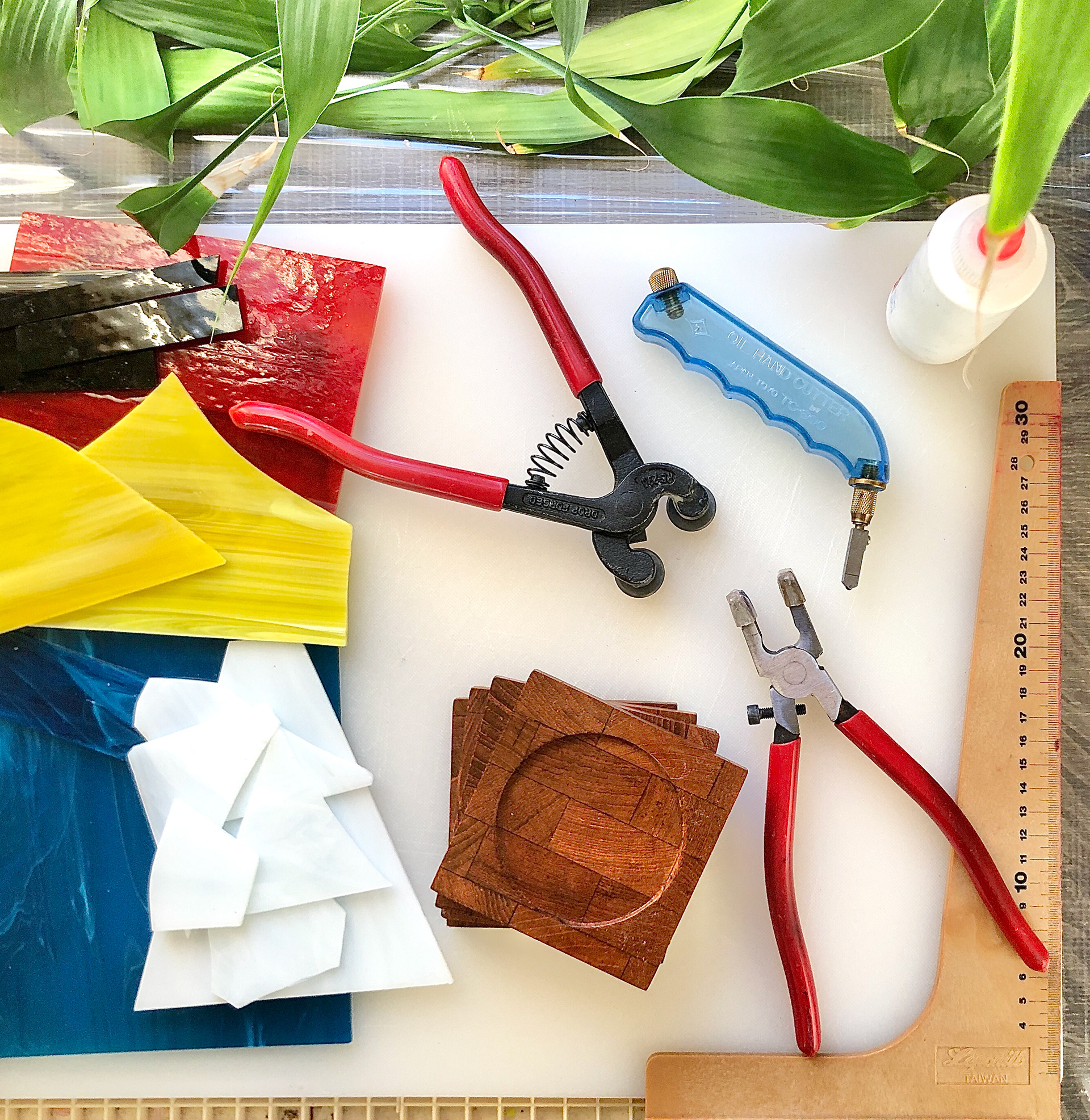 Coloured glass sheets, glass cutting and scoring tools, glue, a ruler and the coasters sit on a cutting board, ready to be used.
