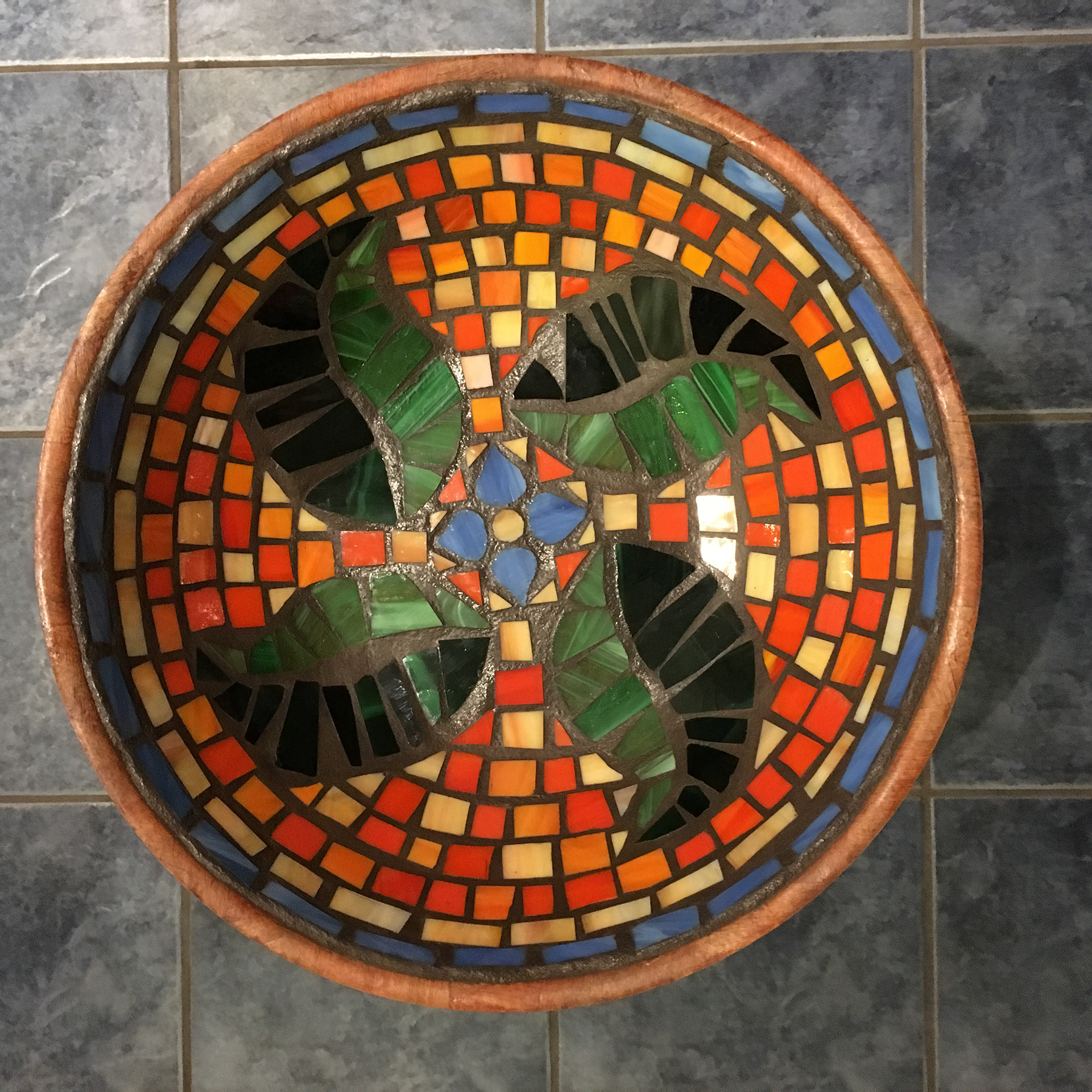 Finished fruit bowl on the floor with mosaics. The design includes a blue flower in the middle, with 4 green leaves surrounding it. The background is covered in square, orange tiles and framed by a light blue rim.