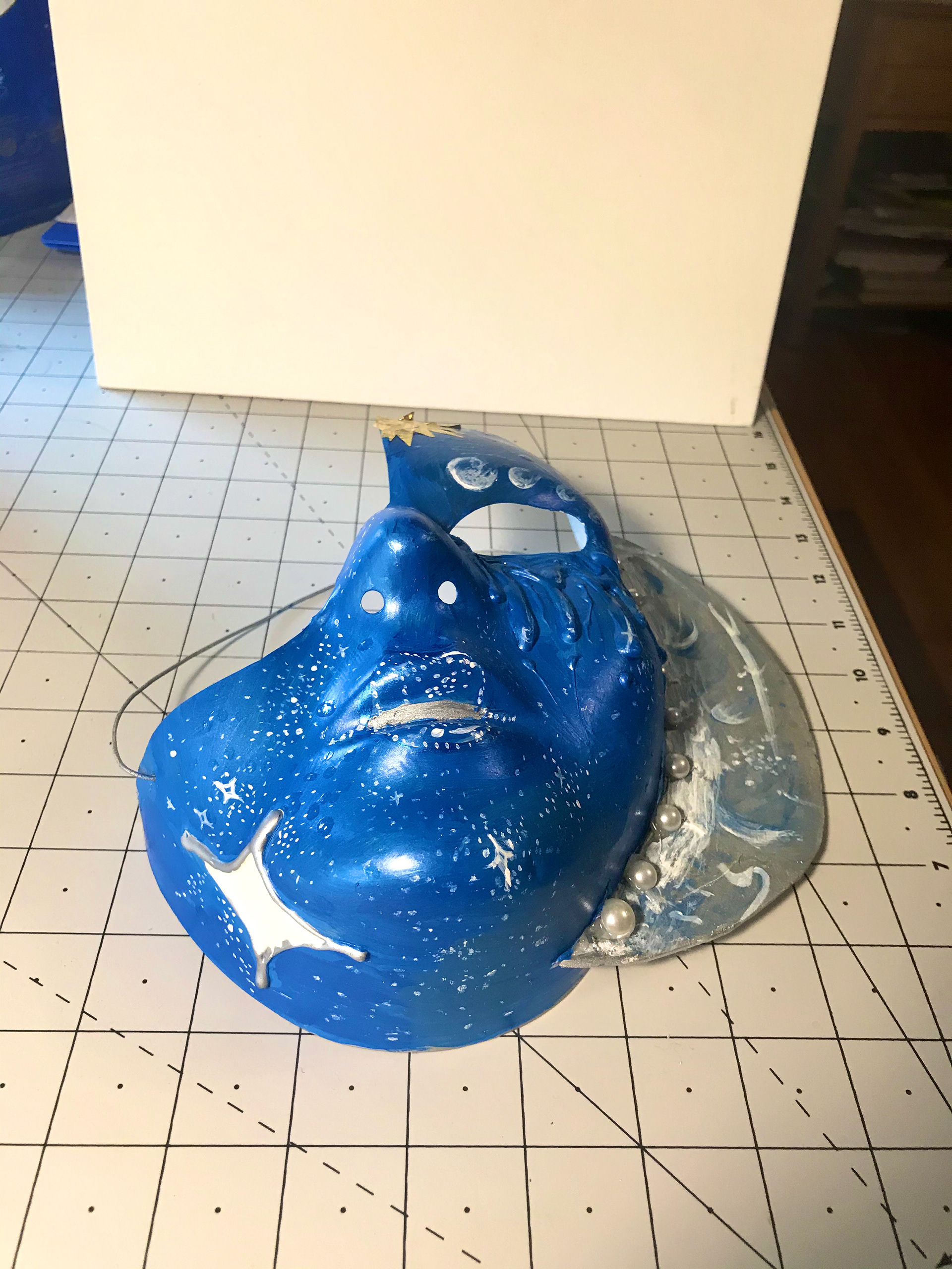 3. Using white and silver paint, I dotted tiny stars and swirls into the blue of the mask. Across the eyebrow I painted phases of the moon. On the crescent moon on the side of the face I painted craters with blue highlights and added plastic pearls to blend the foam into the mask, also imitating the moon phases on the eyebrow.