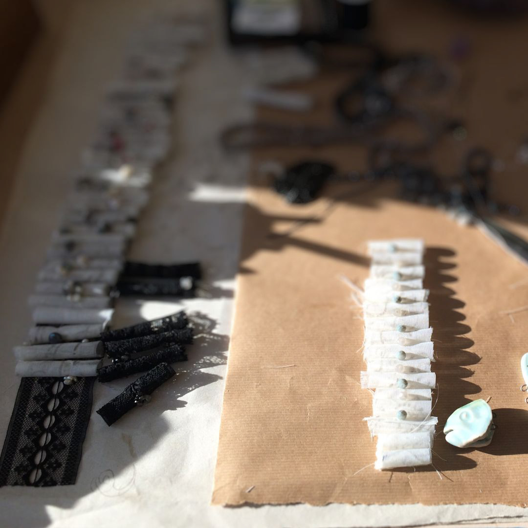 work in progress image artist studio showing two long rows of fabric scrolls on a sunlit table with beads and pearls and laces 
