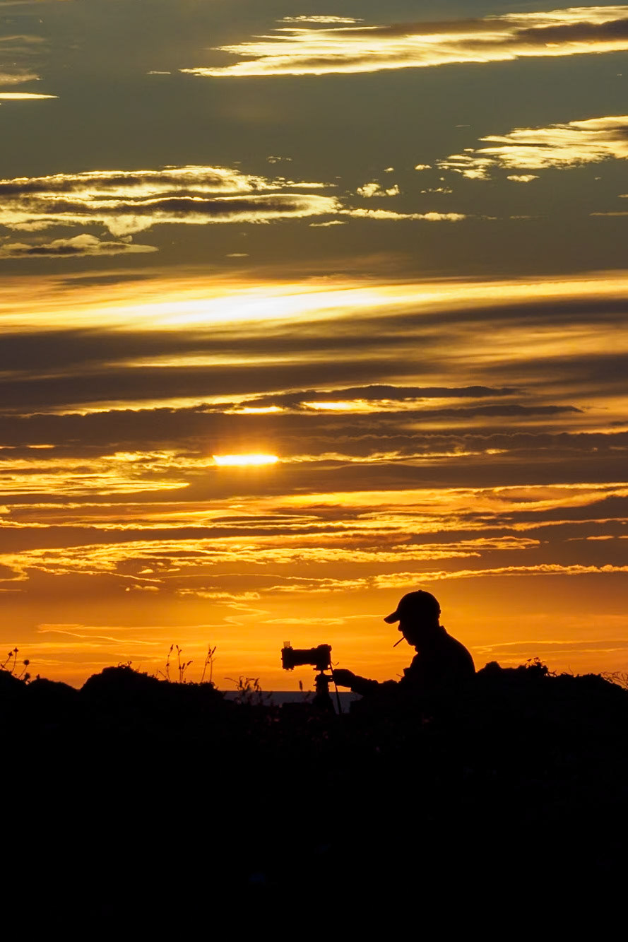 A photographer is silhouetted against the sunset sky, photographing the scene too. Somewhere in Menorca, Spain.