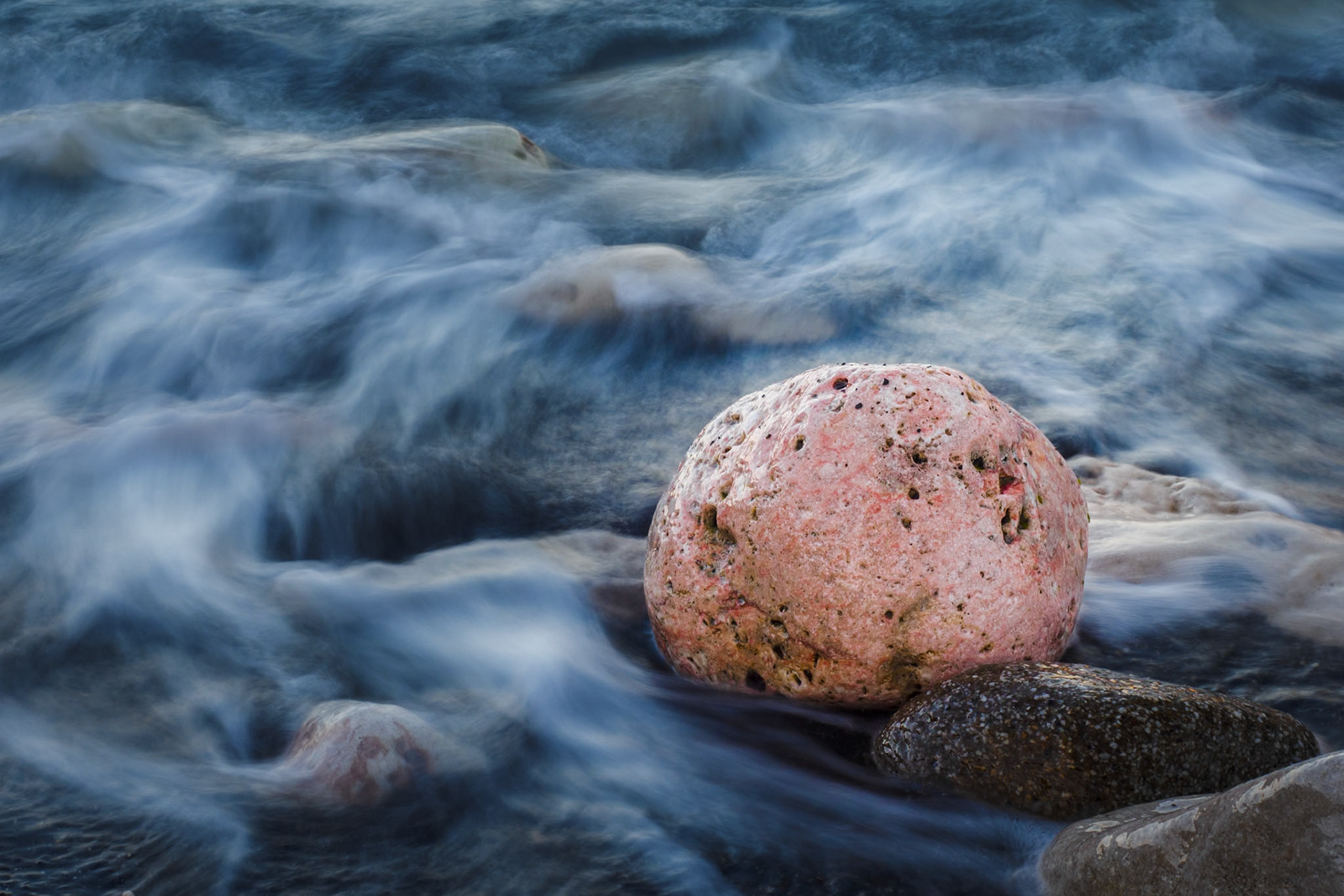 A pink rock stands in contrast with the dark pebbles on the beach, as the sea water swirls around it. Shot taken near the Mola Fortress (Fortalesa de la Mola) in Menorca.