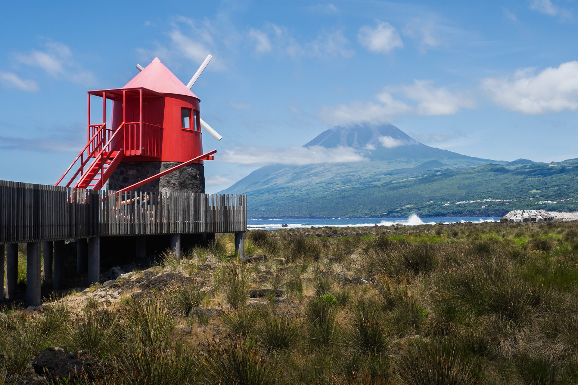 A striking red windmill with a wooden walkway overlooks the ocean, with the majestic Mount Pico rising in the background on Pico Island in the Azores, Portugal.