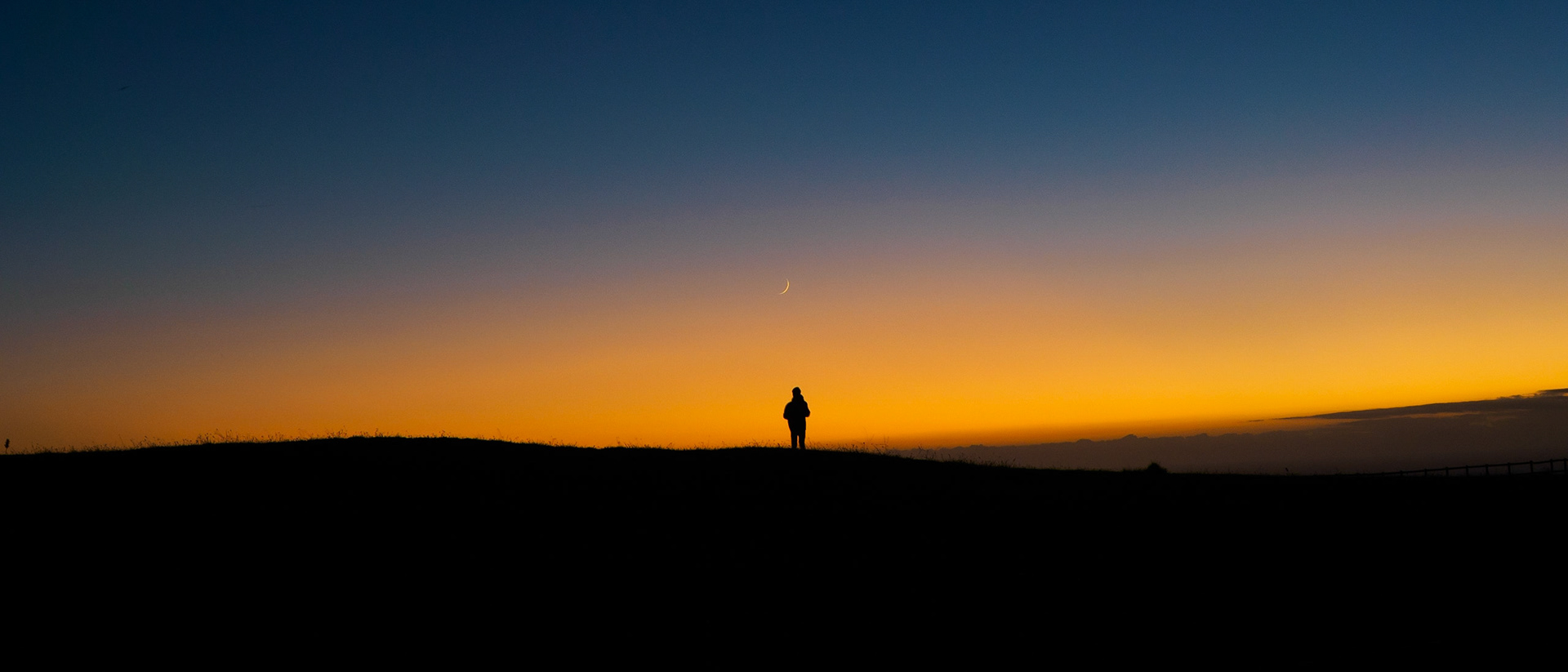 Sillouhette of a man standing directly beneath a waxing crescent moon, at sunset.