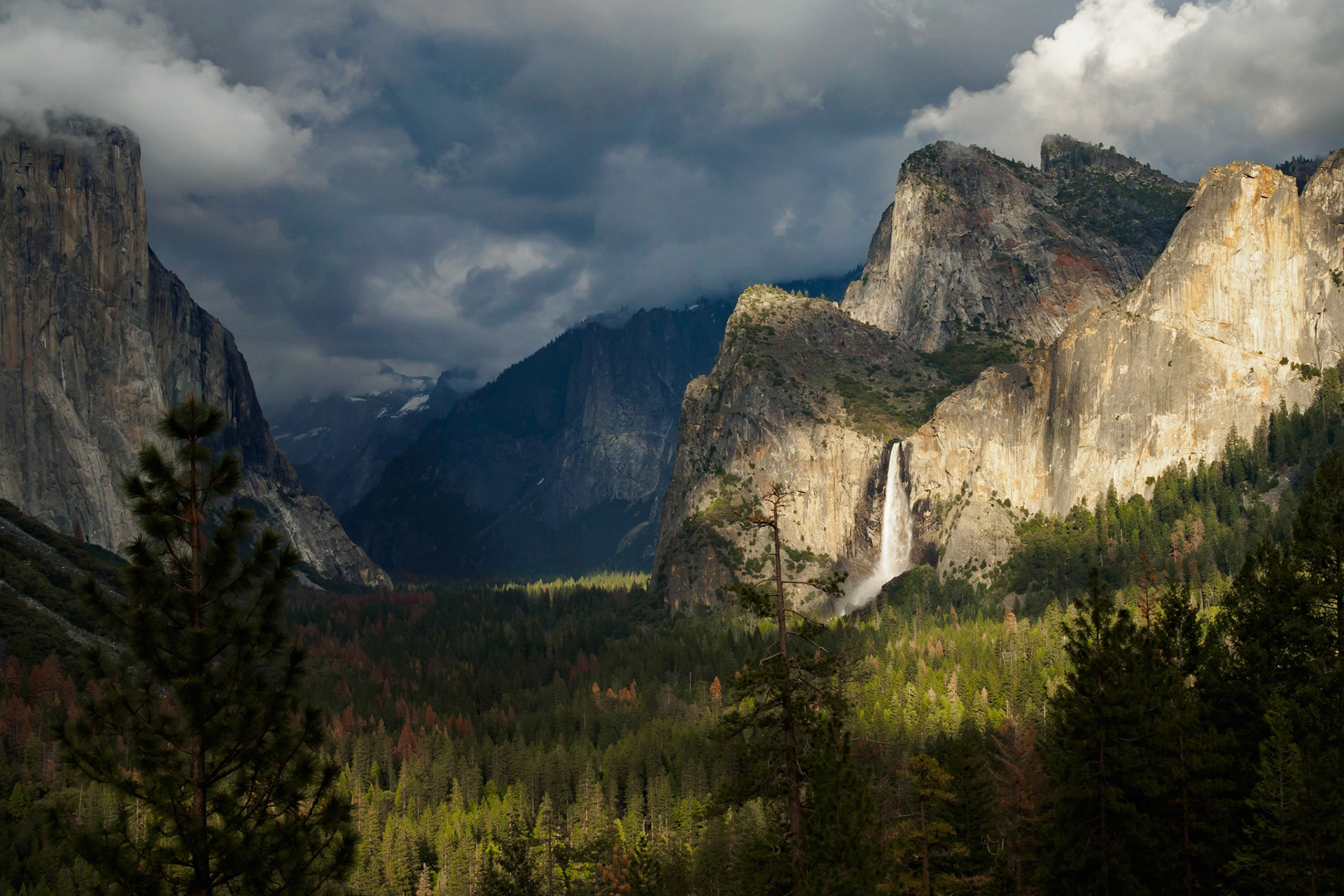 Wide angle view of Yosemite Valley at dusk, wtih overcast skies, sunlight piercing through and illuminating the rocks and Bridalveil Fall