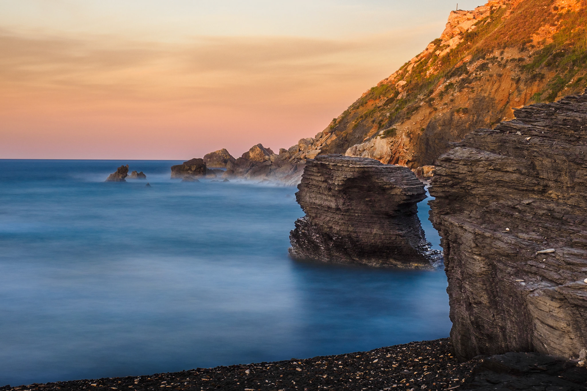 On a pebble beach near the Mola Fortress(Fortalesa de la Mola) in Menorca. I noticed this seastack having a silhouette almost matching the giantrock on the beach itself. I composed the shot so thatthe two edges almost line up, with a sliver of seavisible to separate the two halves. Meanwhile thesetting sun illuminates the cliff with a beautiful goldenlight.