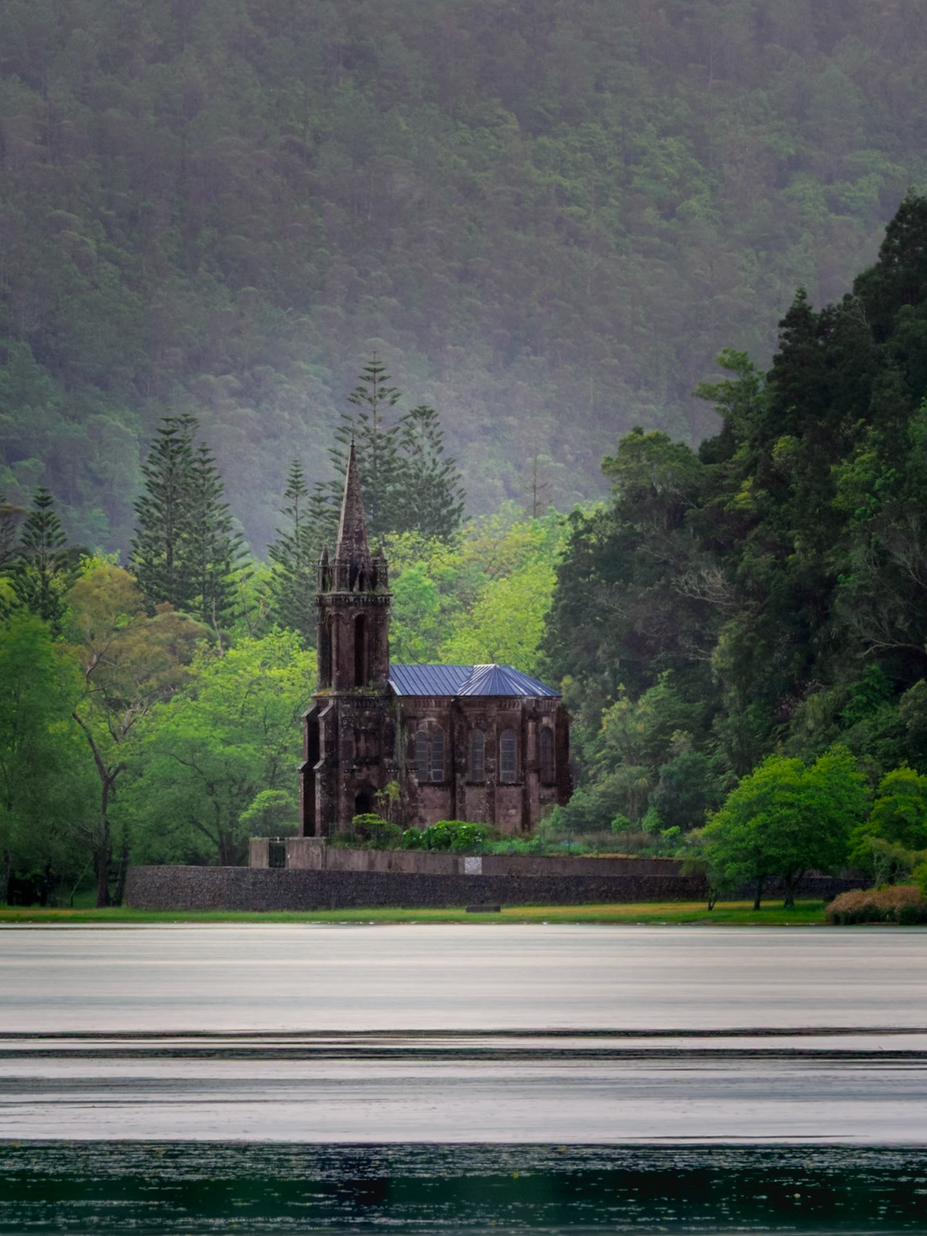 The Chapel of Nossa Senhora das Vitórias, a striking Gothic Revival structure, situated on the tranquil shores of Furnas Lake (Lagoa das Furnas) on São Miguel Island. It stands strong on the edge of the crater lake, the dark stone in stark contrast to the lush green foliage around it.