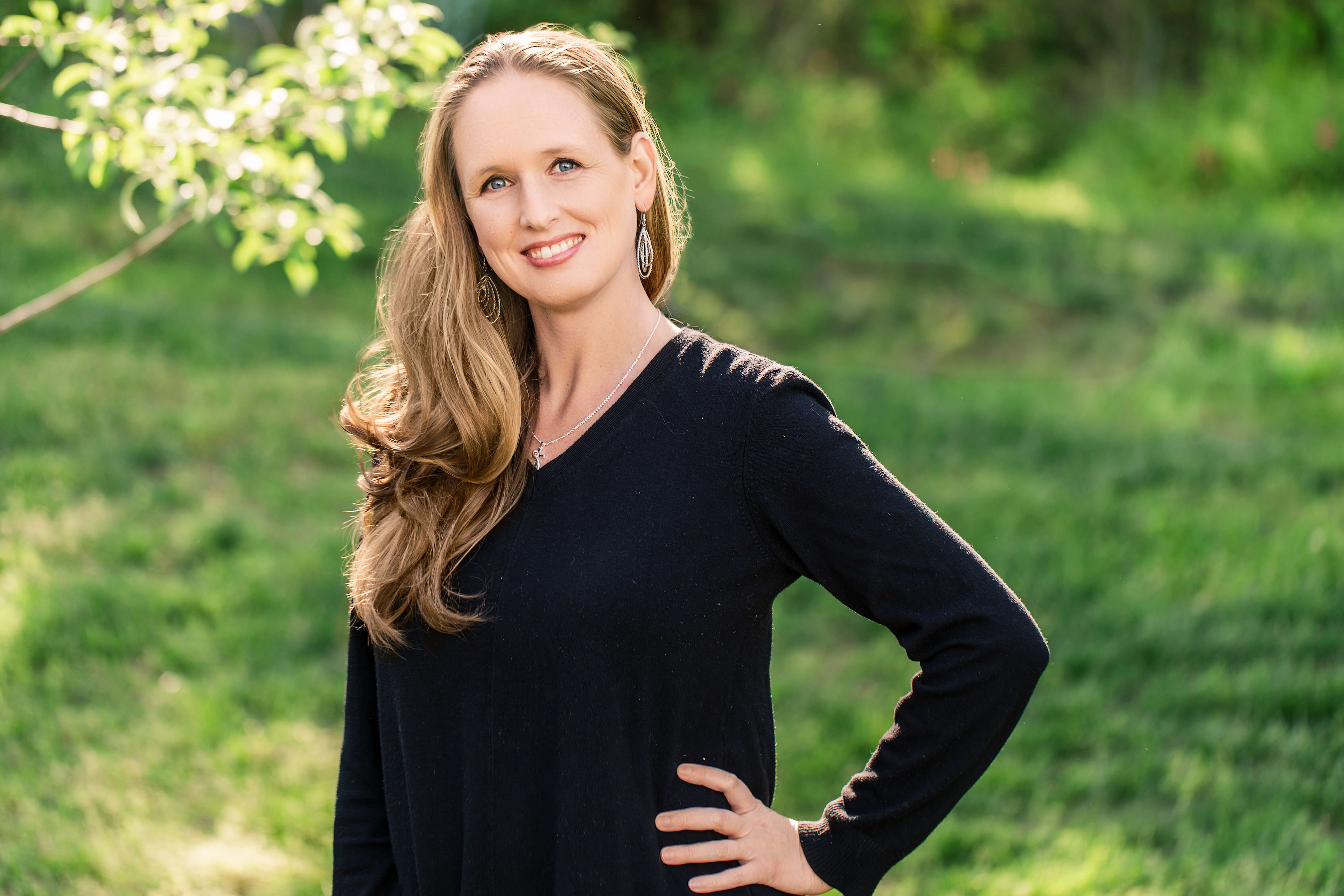 Photo of Dianne Greene with a genuine, sweet, and caring smile surrounded by grass and a glowing tree branch. She is the main photographer and owner of Remembering Stones Photography,