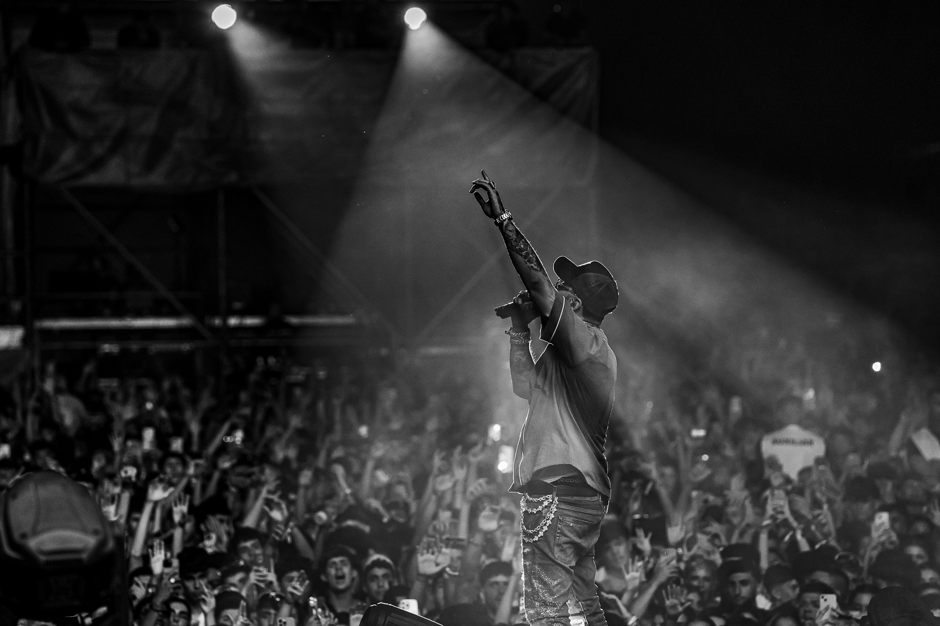 Anuel AA, uno de los máximos exponentes del trap y reggaetón, actuando en blanco y negro ante una multitud en el Reggaeton Beach Festival Tenerife 2024, irradiando energía sobre el escenario en las Islas Canarias.