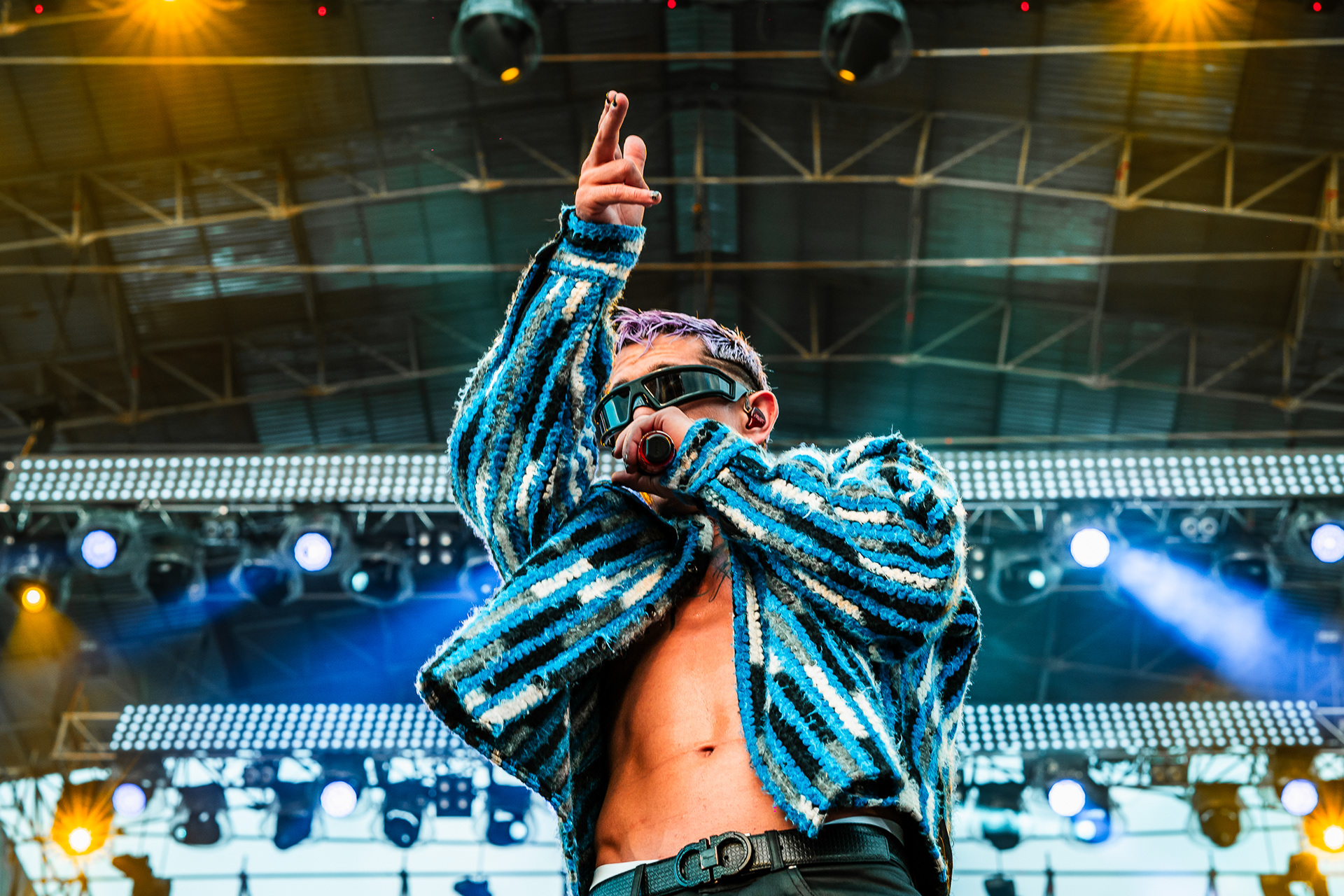 Lenny Tavárez, popular exponente del reggaetón, luciendo chaqueta de rayas y cabello púrpura mientras canta en el Reggaeton Beach Festival Tenerife 2024, rodeado de un gran escenario iluminado y un público entusiasta en las Islas Canarias.